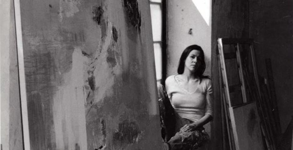 Black-and-white photo of a woman sitting in an art studio beside a large canvas and a leaning ladder by a window.