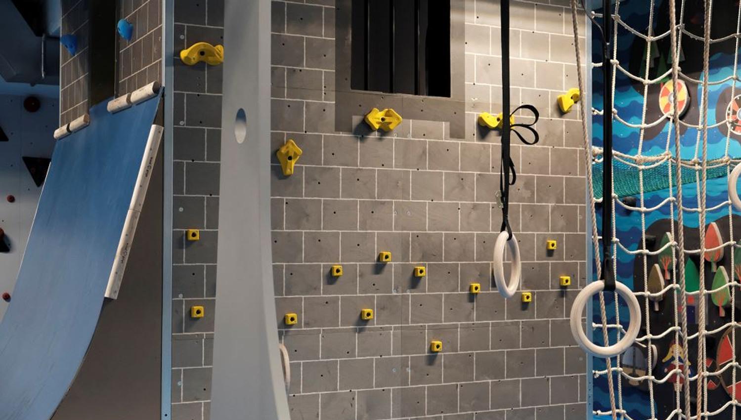Indoor adventure gym with a gray climbing wall, yellow holds, hanging gymnastic rings, and a rope net..
