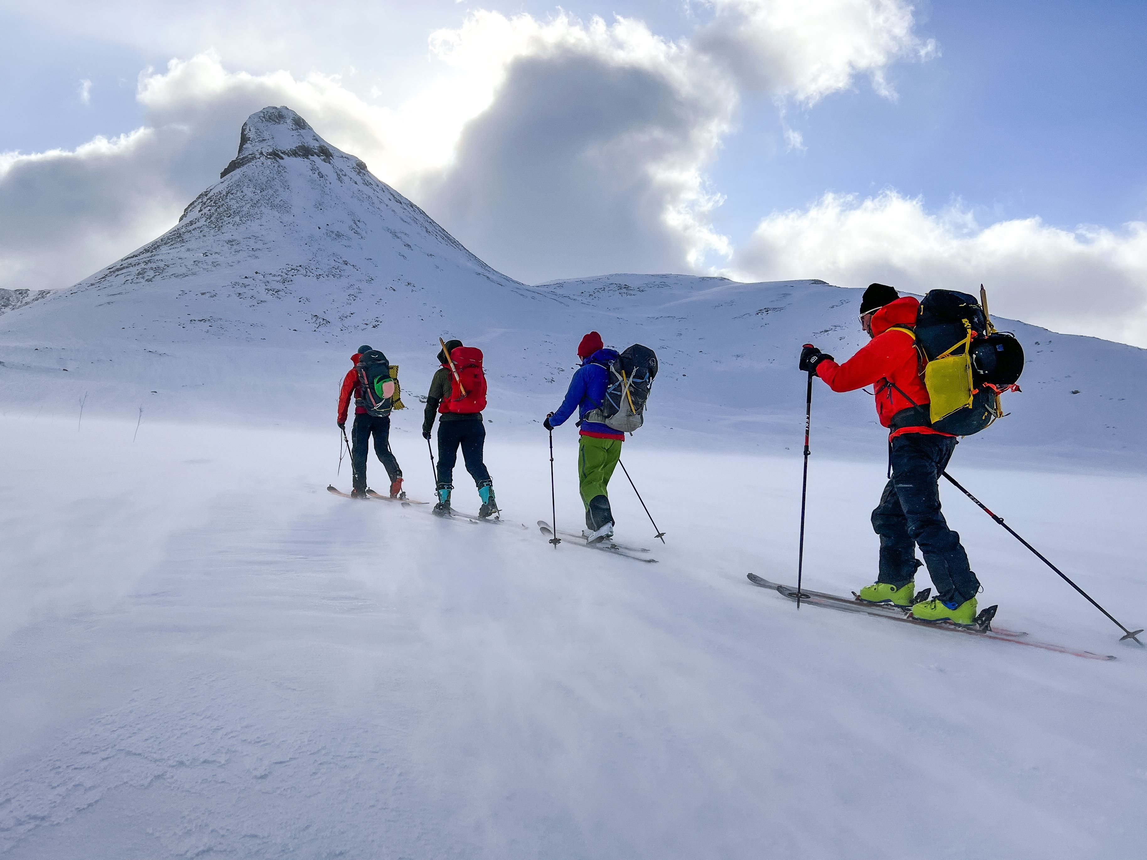 Vier skiërs met rugzakken beklimmen een door de wind geteisterde besneeuwde helling naar een steile berg onder een bewolkte blauwe lucht..