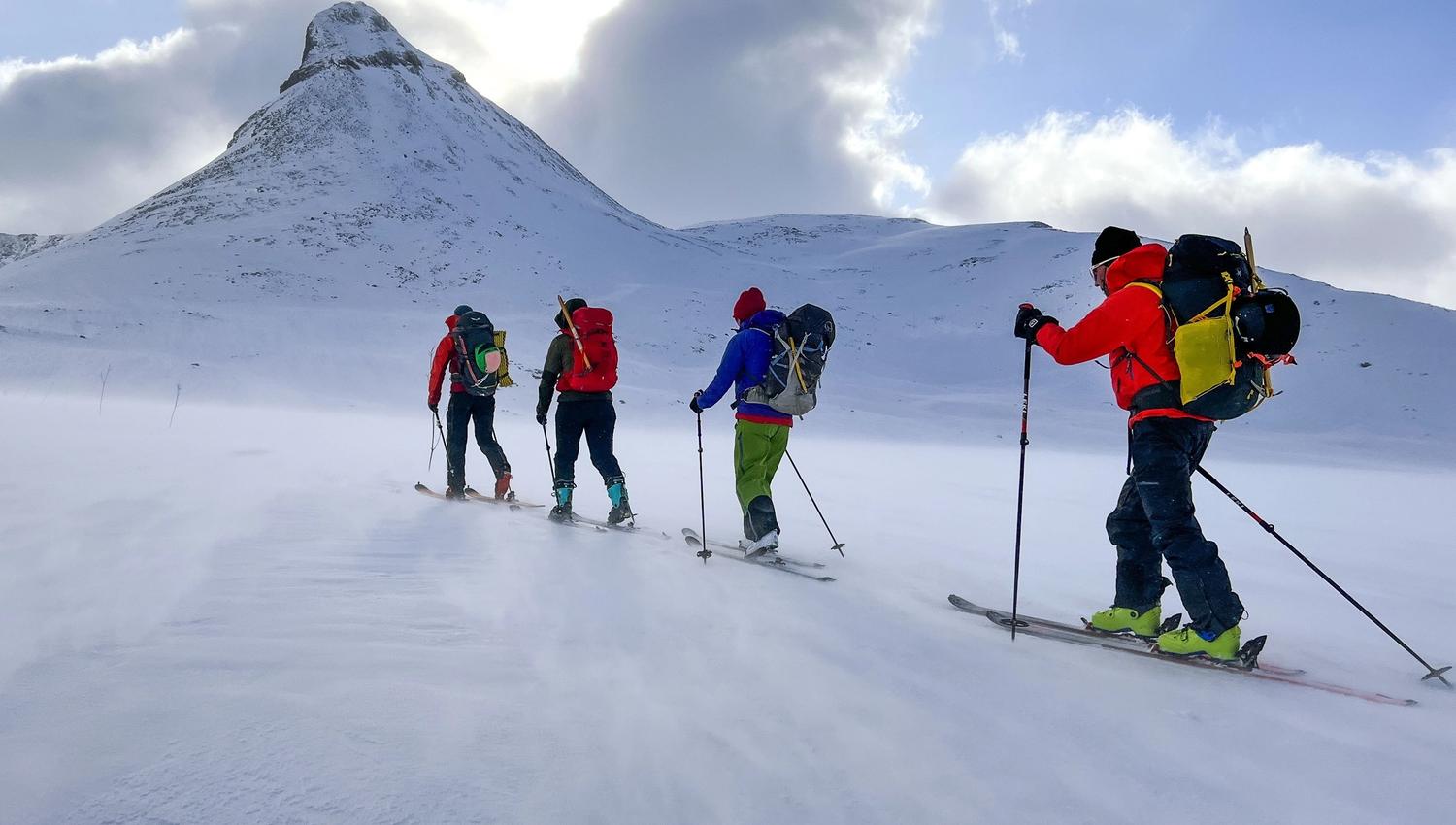 Vier skiërs met rugzakken beklimmen een door de wind geteisterde besneeuwde helling naar een steile berg onder een bewolkte blauwe lucht..