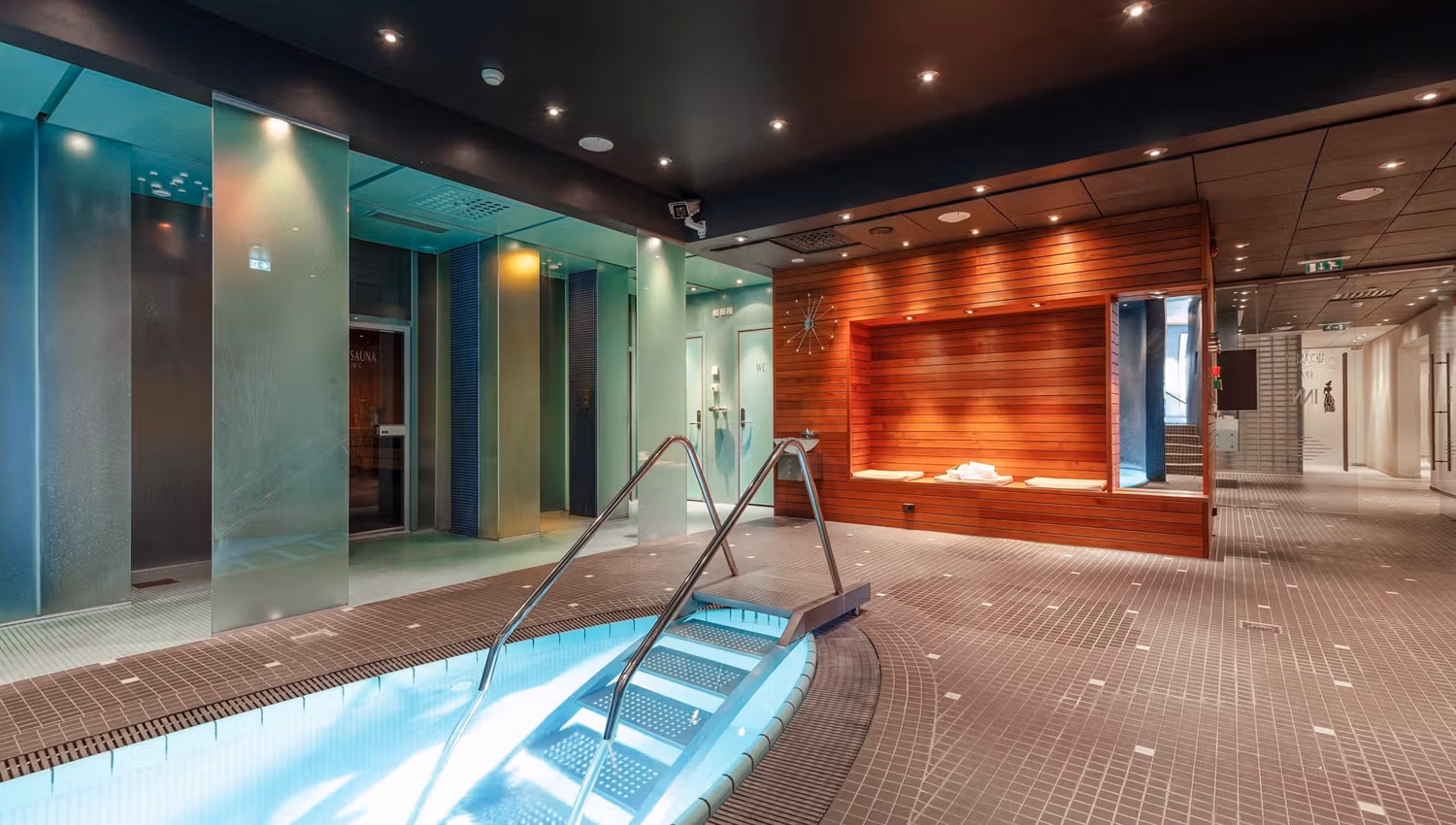 Indoor spa pool edge with metal handrails and lit steps, beside frosted glass walls and a wood bench alcove at Alexandra Spa..