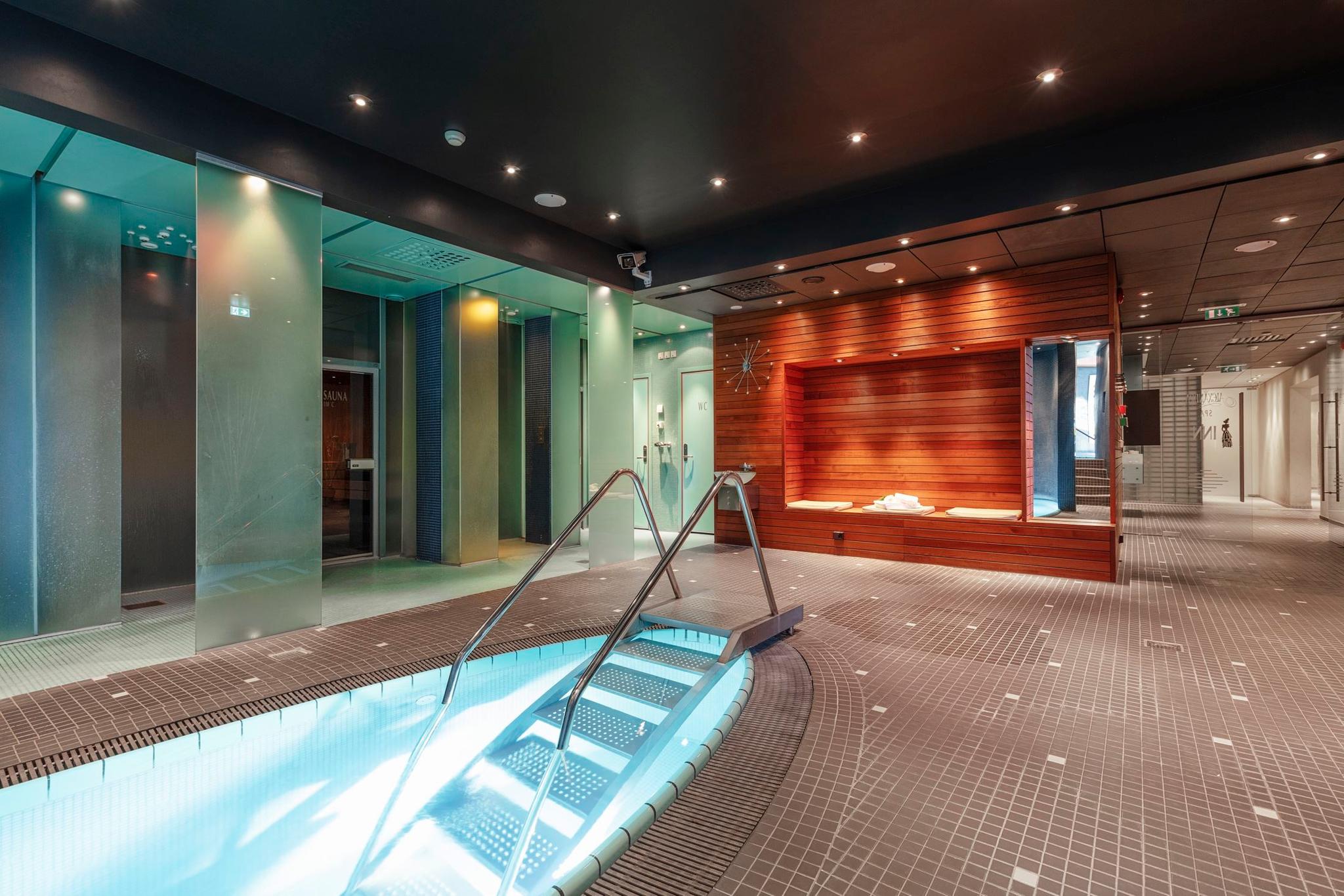 Indoor spa pool edge with metal handrails and lit steps, beside frosted glass walls and a wood bench alcove at Alexandra Spa..
