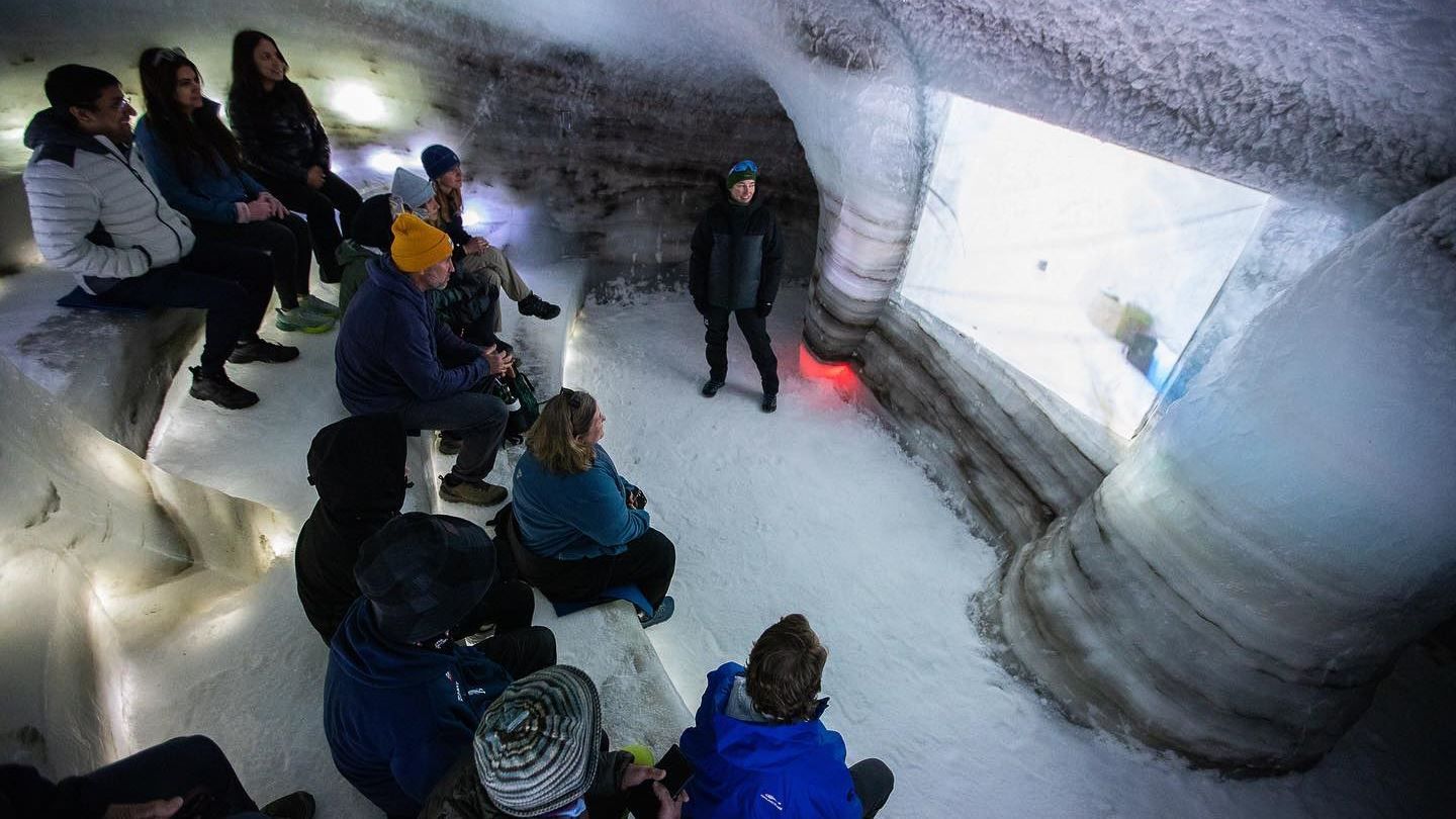 Ei gruppe menneske sit på isbenkar i ei isgrotte og ser mot ein guide ved eit stort opplyst isvindauge..