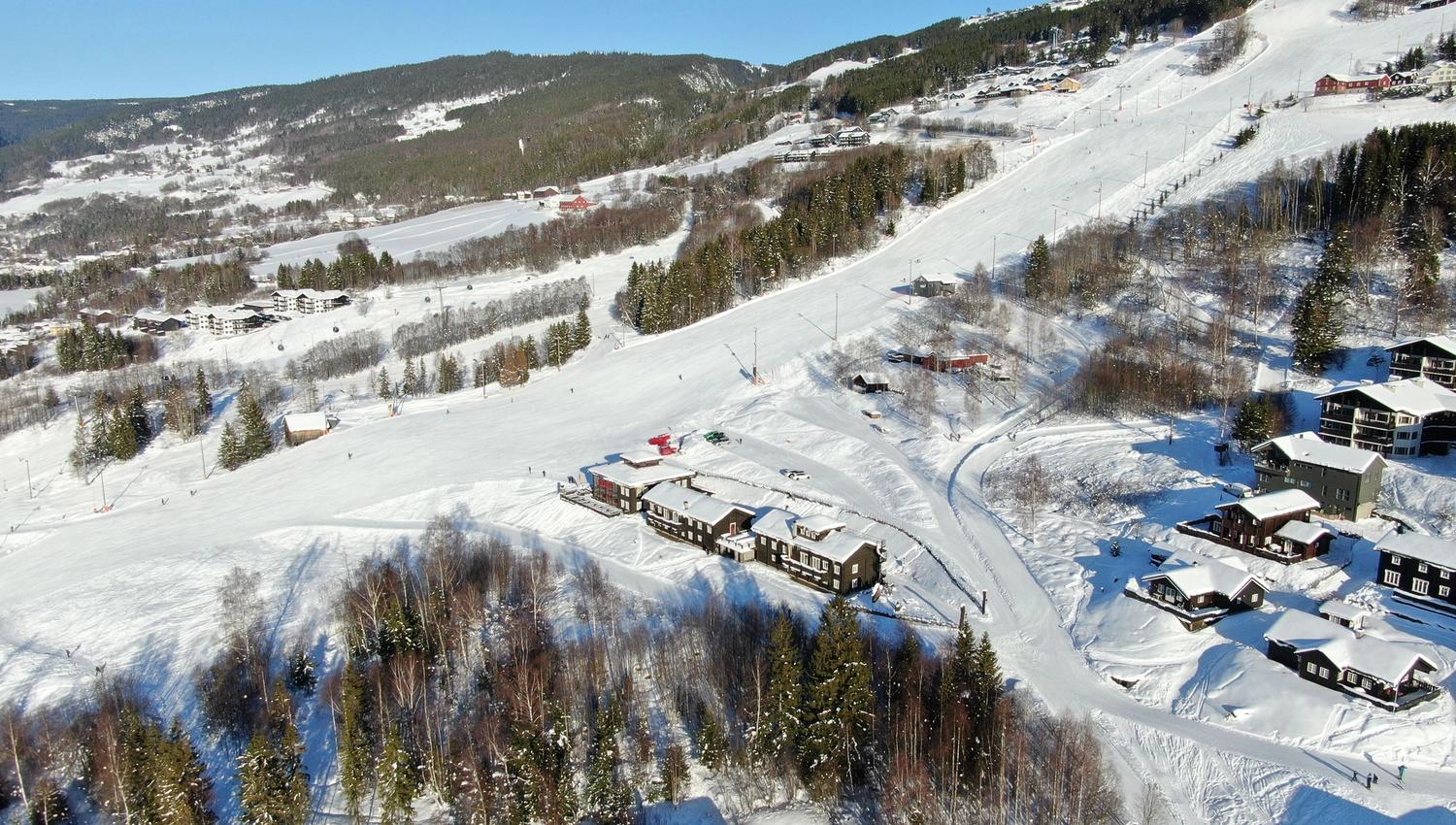 Luftfoto av snødekt skianlegg med brede løyper, hytter og grantrær i fjellsiden under blå himmel..