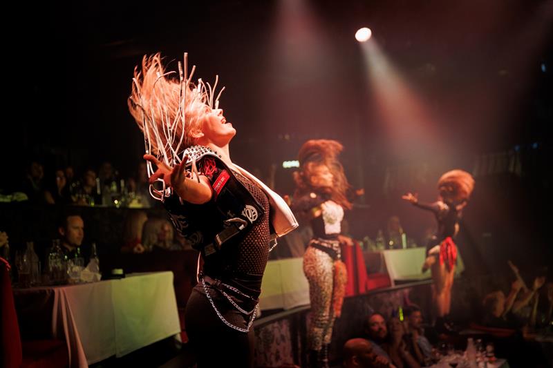 En energisk danser svinger håret i takt med musikken under et intenst sceneshow med lys og publikum tett på. Scenen er fylt med kraft og bevegelse.