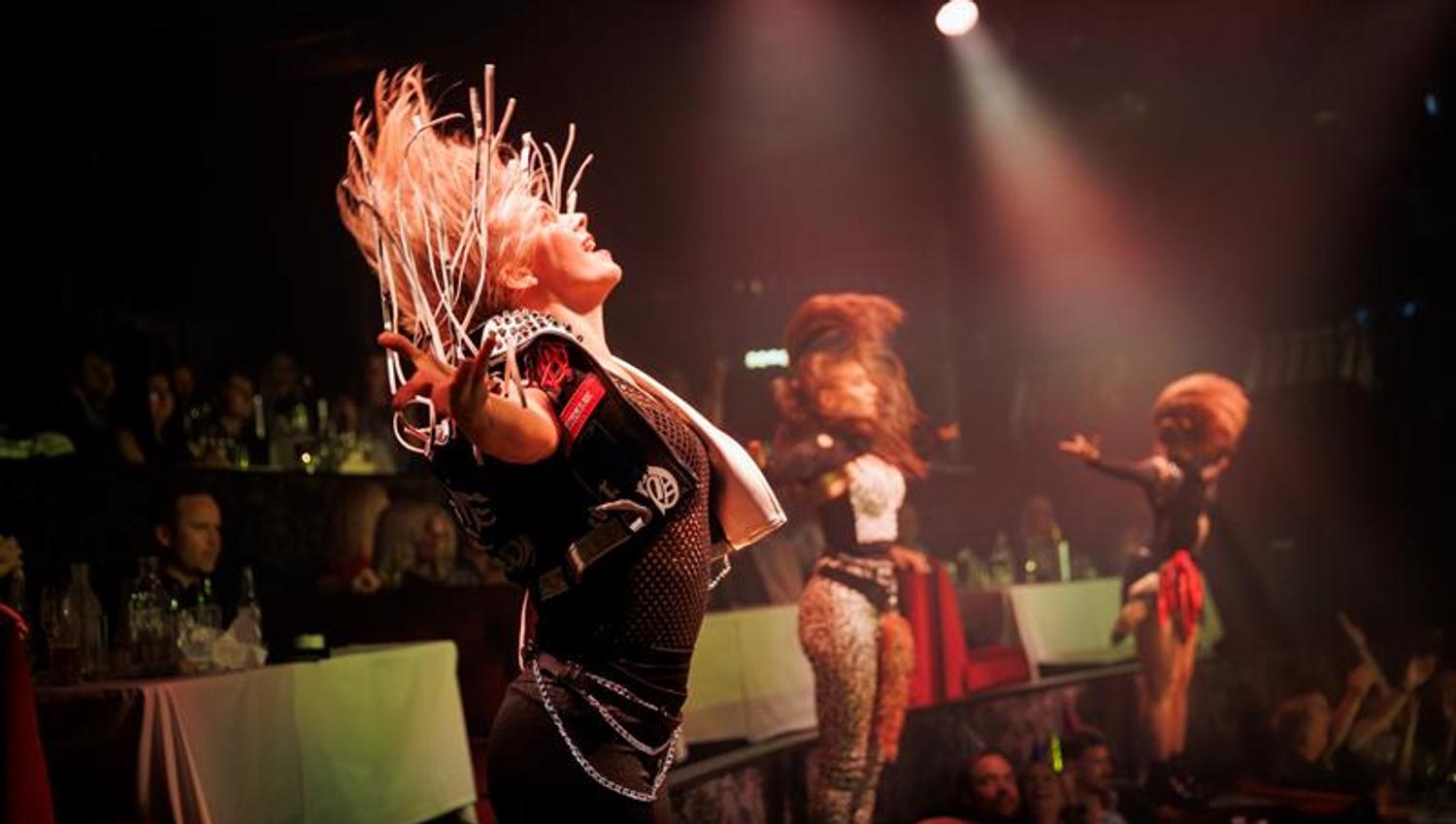 En energisk danser svinger håret i takt med musikken under et intenst sceneshow med lys og publikum tett på. Scenen er fylt med kraft og bevegelse.
