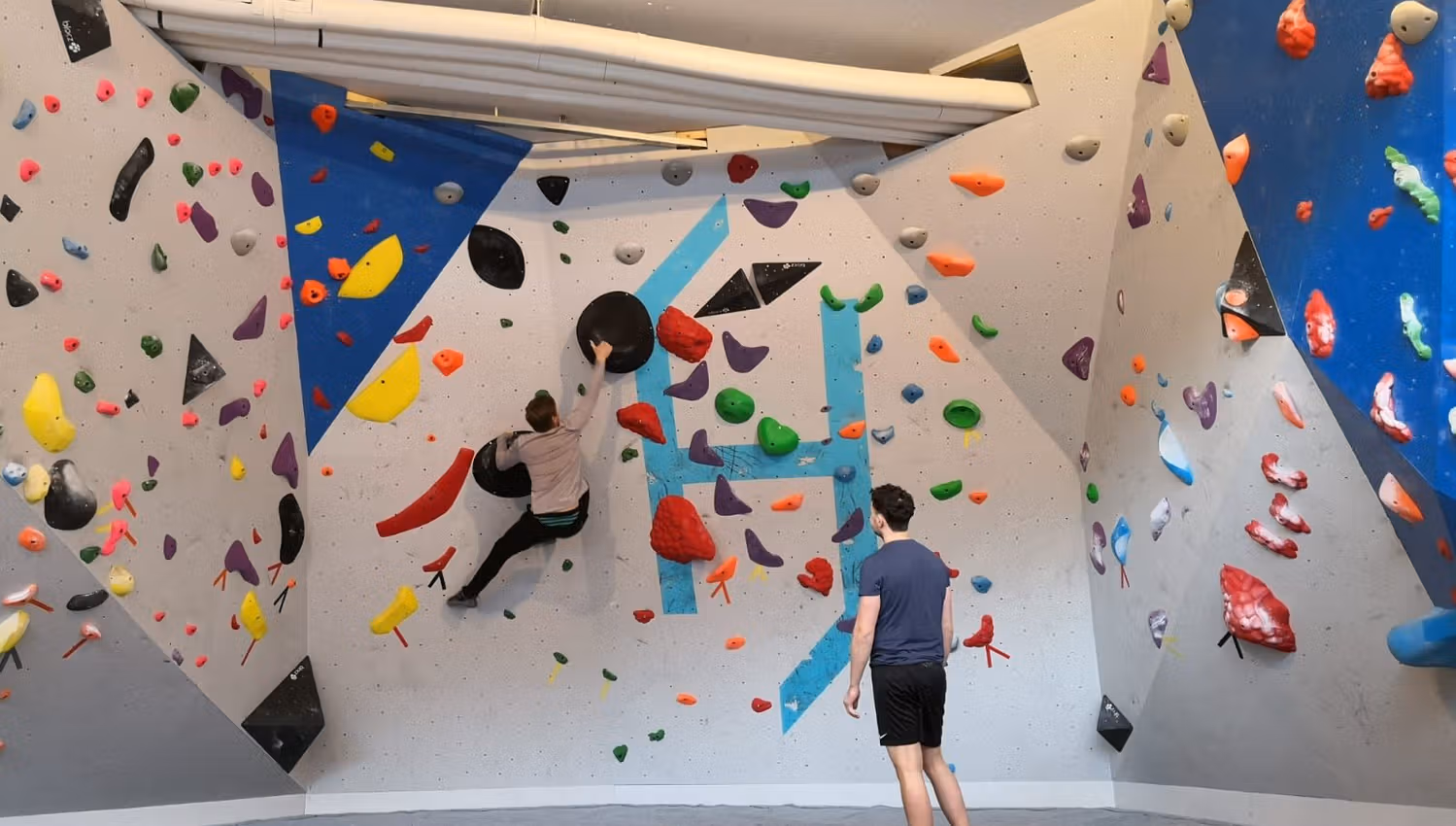 Two people in a bouldering gym; one climbs on a colorful wall of holds while another stands watching below..
