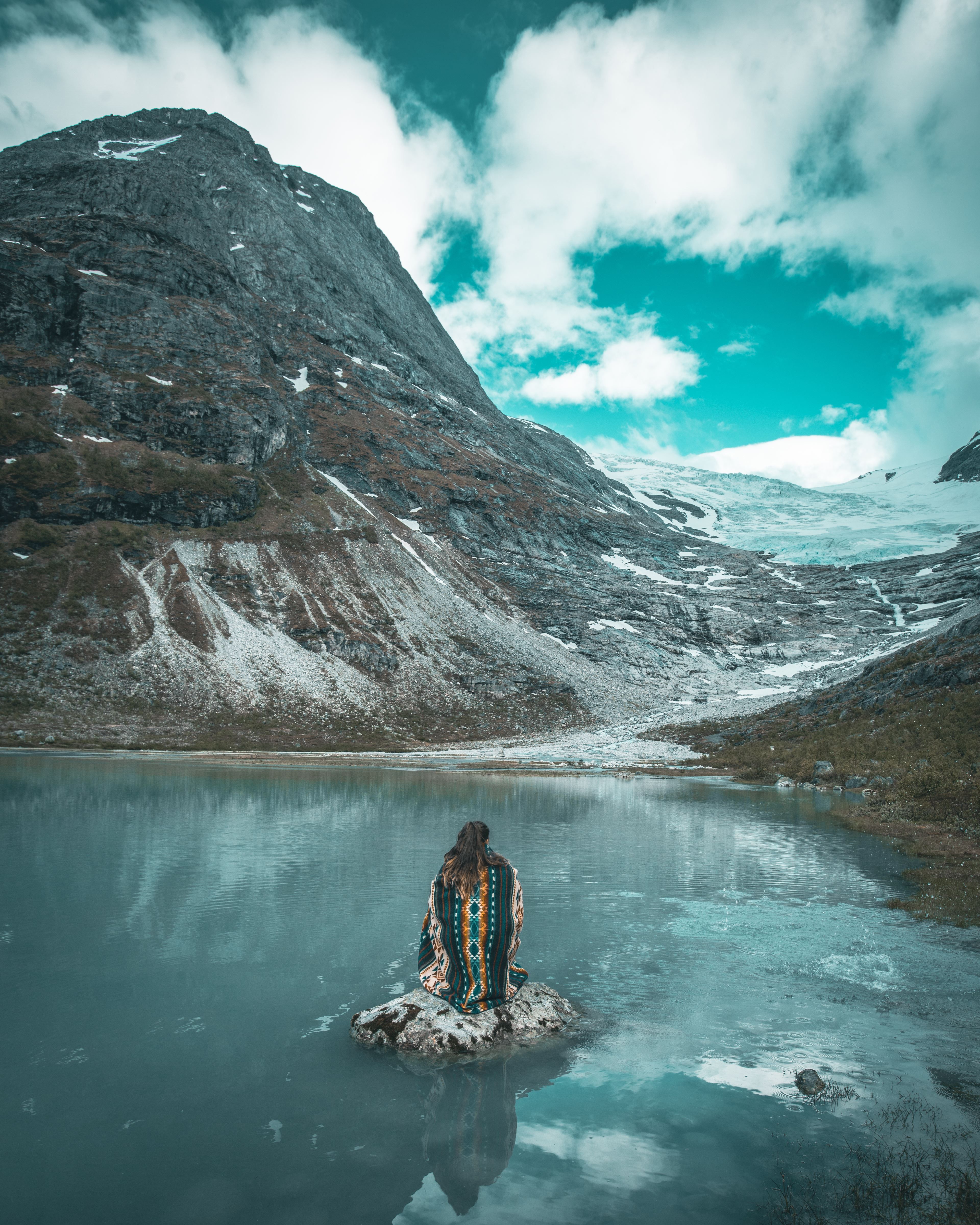 A person sits on a rock in a calm lake, facing rugged mountains with patches of snow under a cloudy sky.