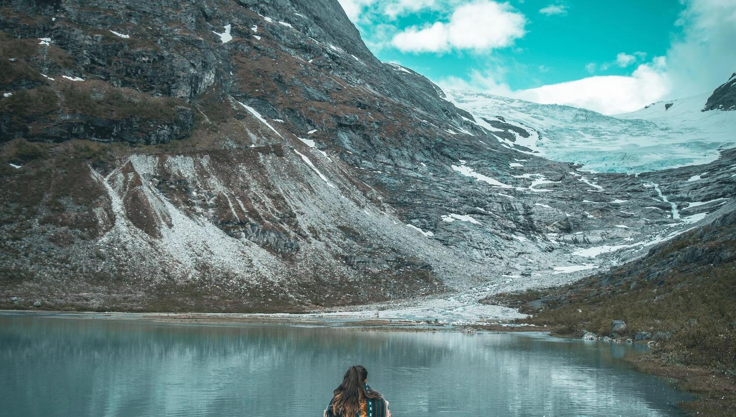 A person sits on a rock in a calm lake, facing rugged mountains with patches of snow under a cloudy sky.