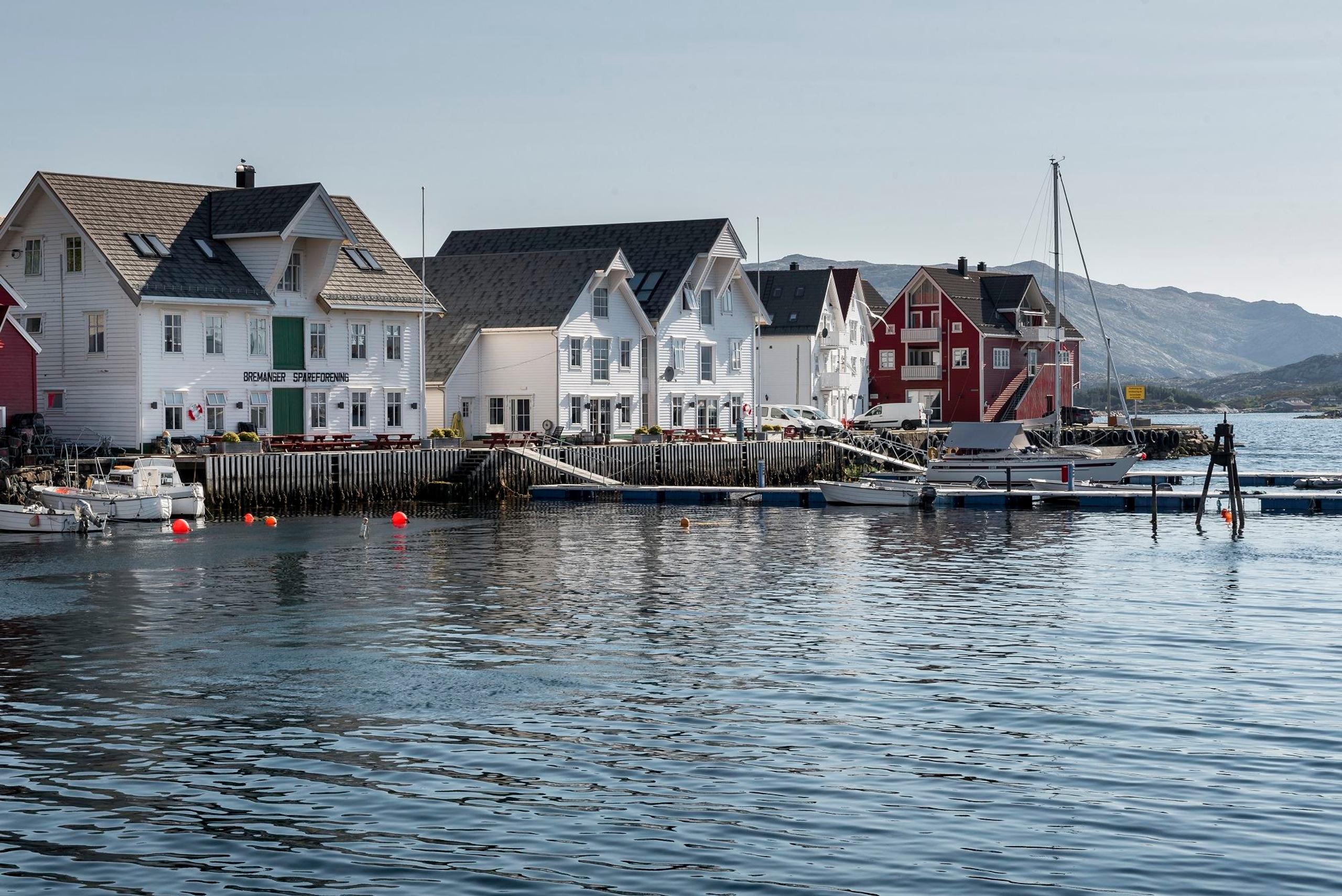 Colorful wooden waterfront houses line a calm harbor with small boats and a sailboat, mountains in the distance.