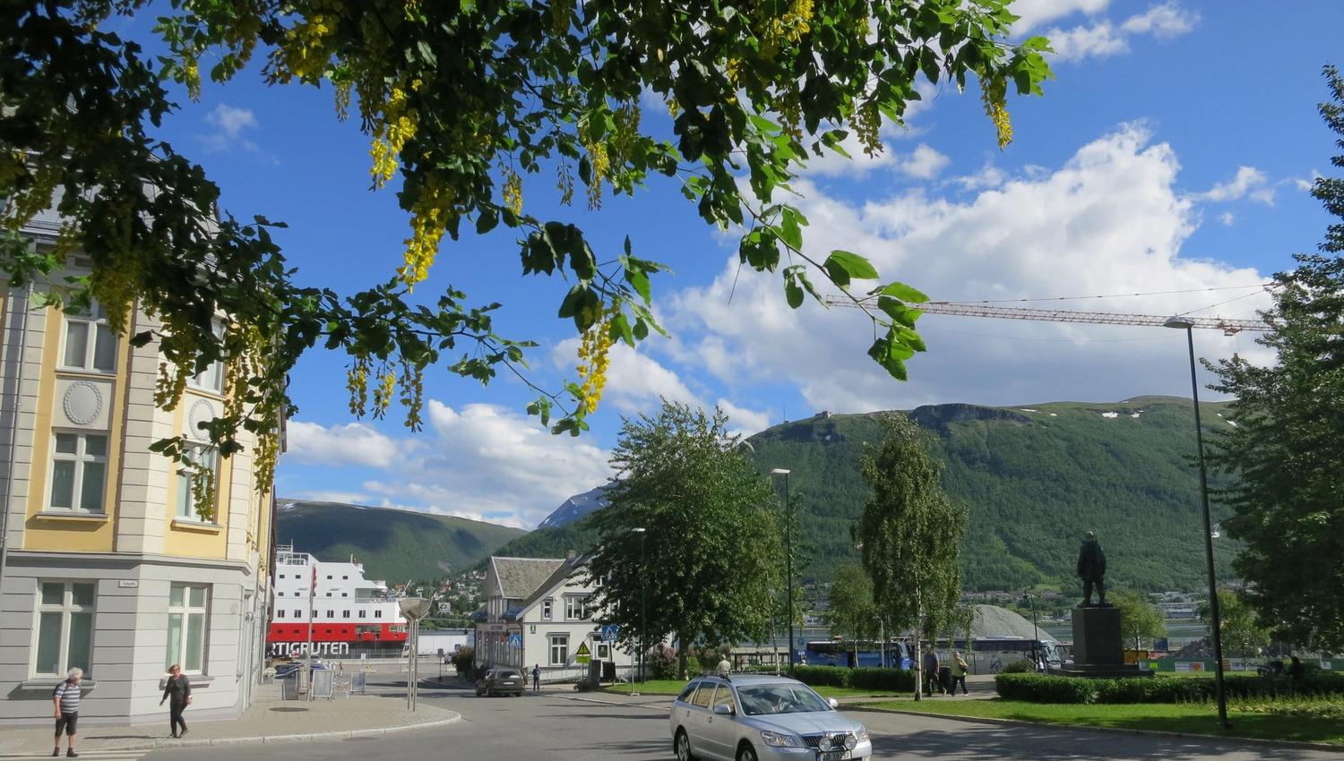 Street scene in Tromsø with people, a car, blooming trees, and mountains in the background.
