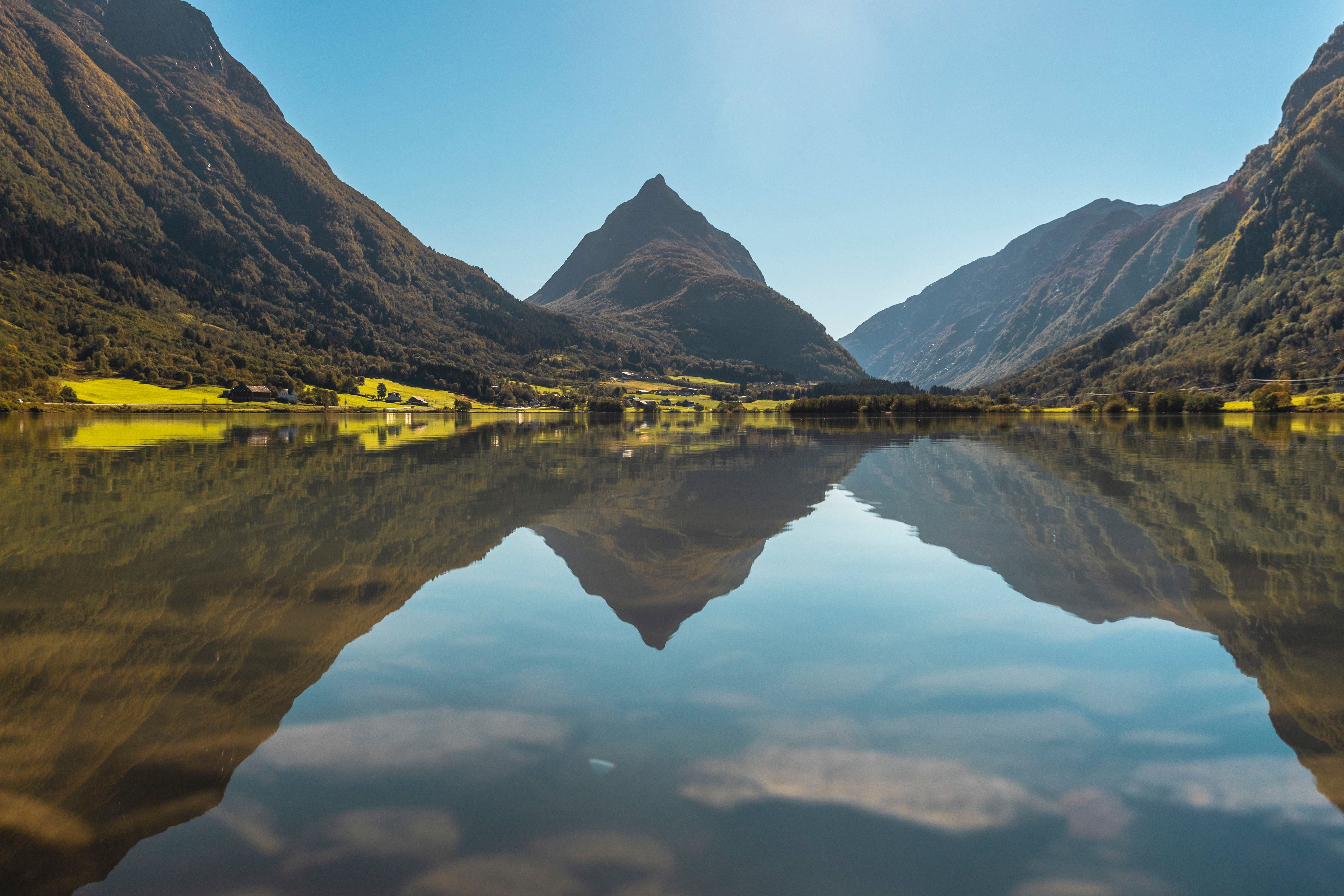 A sharp mountain peak rises between forested slopes, reflected in a still lake under a clear blue sky..
