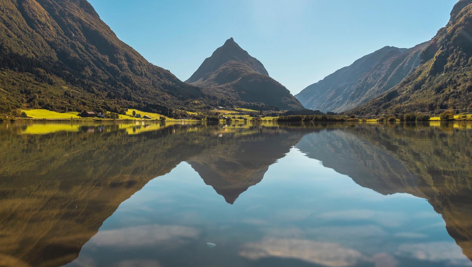 A sharp mountain peak rises between forested slopes, reflected in a still lake under a clear blue sky..