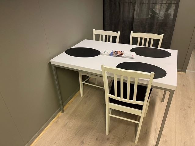 White dining table with three black round placemats, three chairs, and magazines on top in a small room.