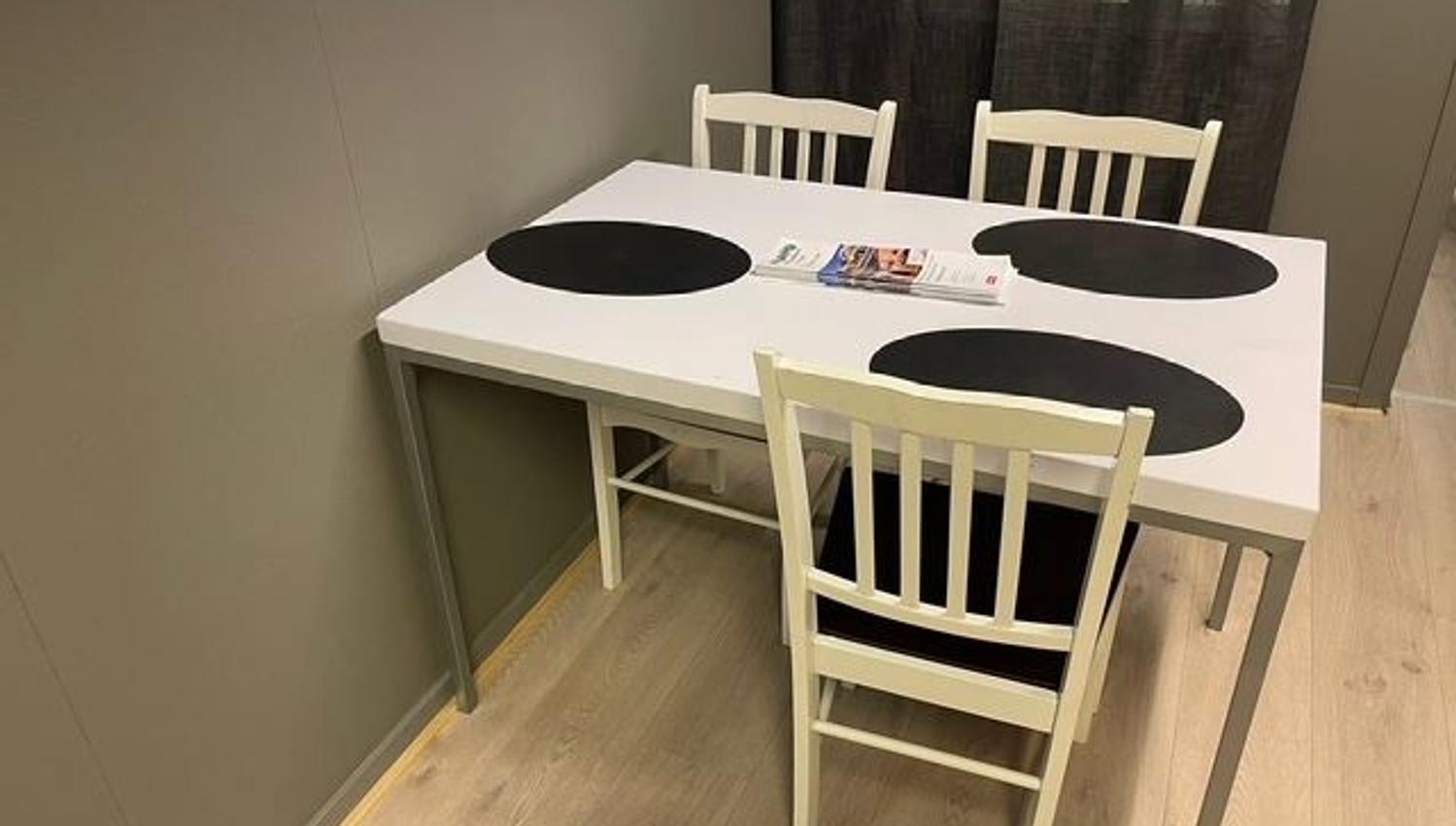 White dining table with three black round placemats, three chairs, and magazines on top in a small room.