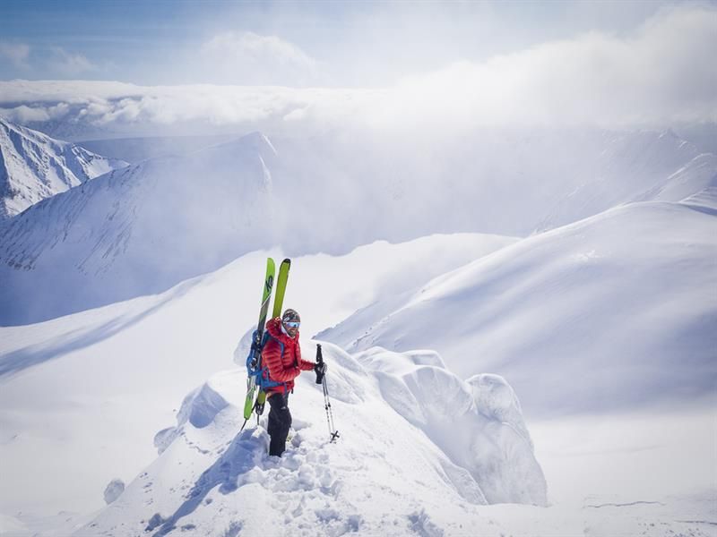 En person står på en fjellrygg med ski i sekken med et fjellanskap i sol i bakgrunnen