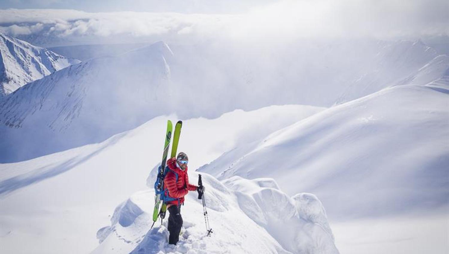 En person står på en fjellrygg med ski i sekken med et fjellanskap i sol i bakgrunnen