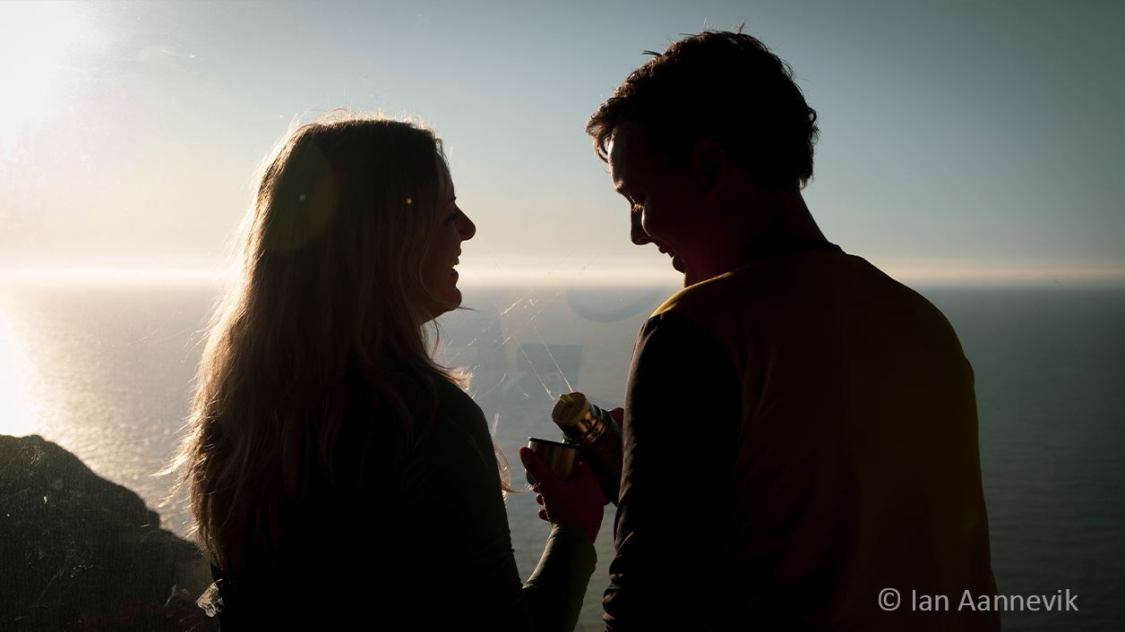 Silhouetted couple facing each other by the sea at sunset, holding a thermos and cup near a coastal overlook..