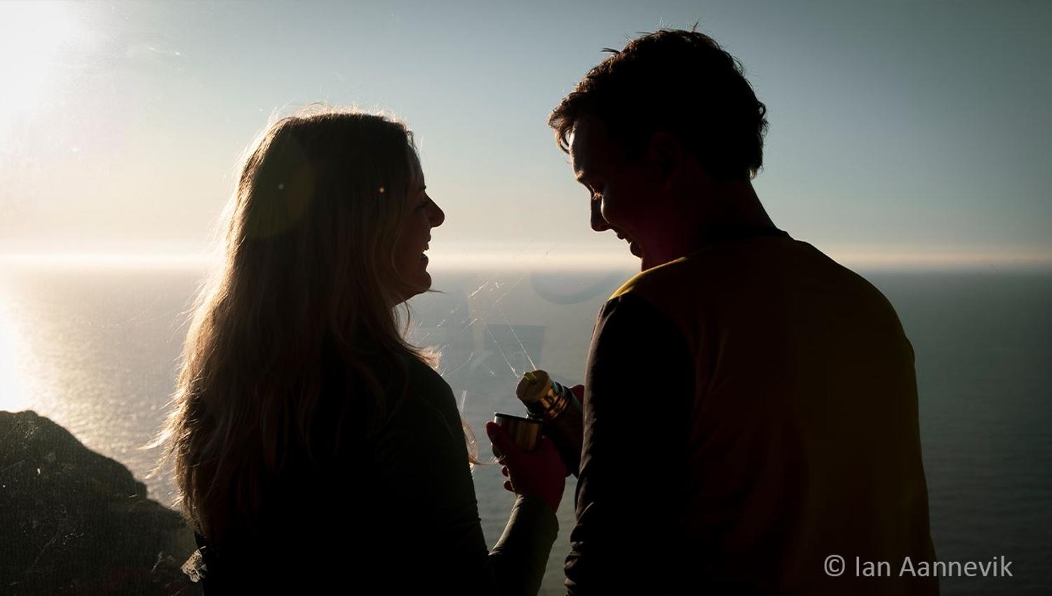 Silhouetted couple facing each other by the sea at sunset, holding a thermos and cup near a coastal overlook..