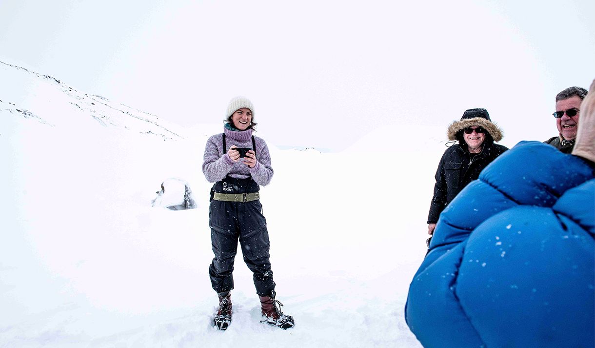 En guide som holder en kopp med varm drikke og prater med gjester i snødekte omgivelser på en isbre