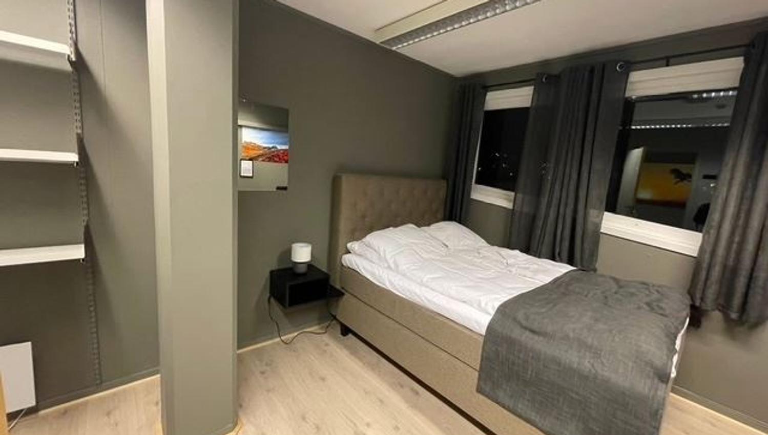 Minimalist bedroom with a single bed, gray walls, built-in shelves, nightstand lamp, and curtained windows.