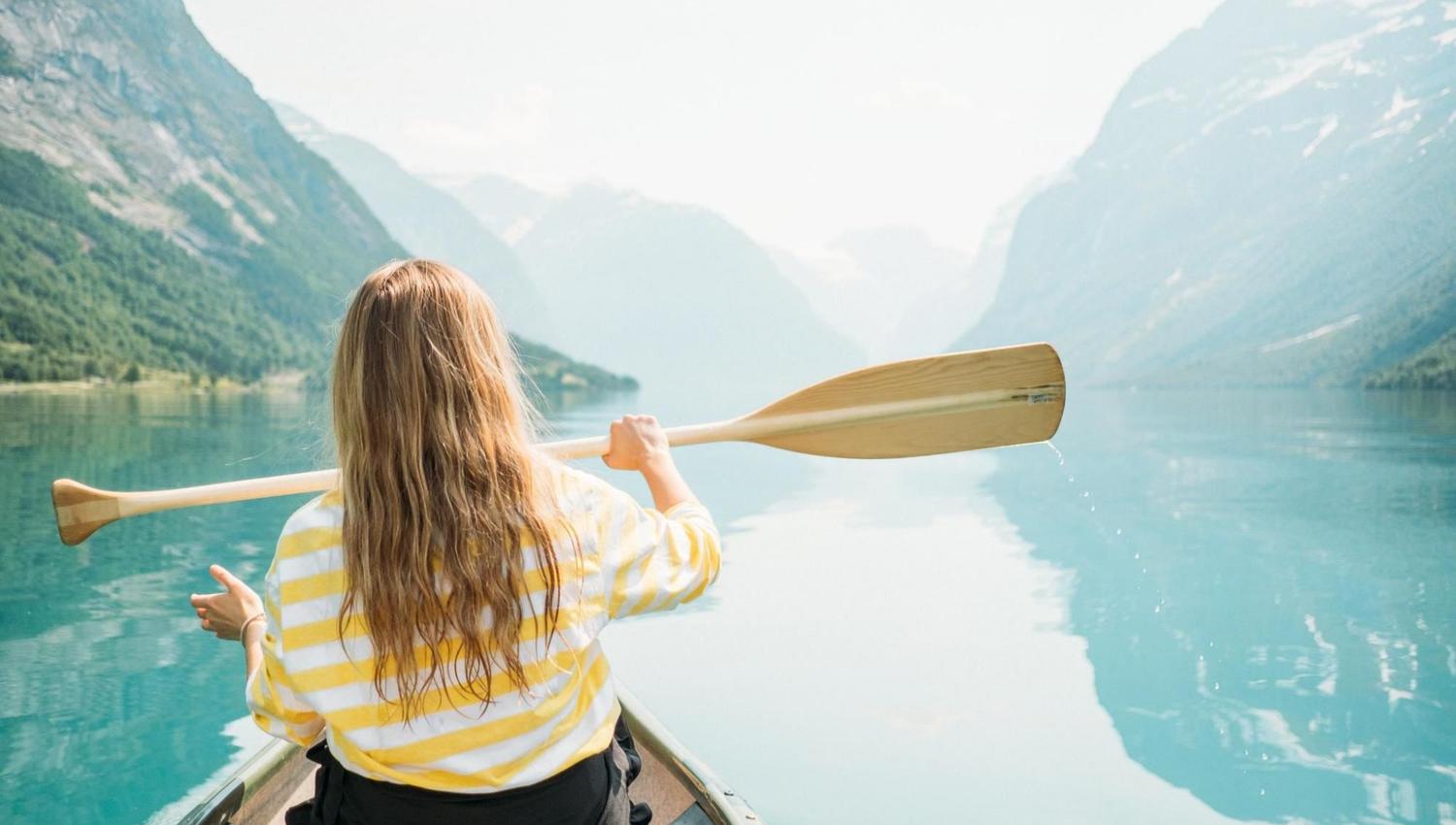 Rear view of a person paddling a canoe on a turquoise Lovatnet lake, surrounded by steep green mountains and haze in Lodalen Valley..