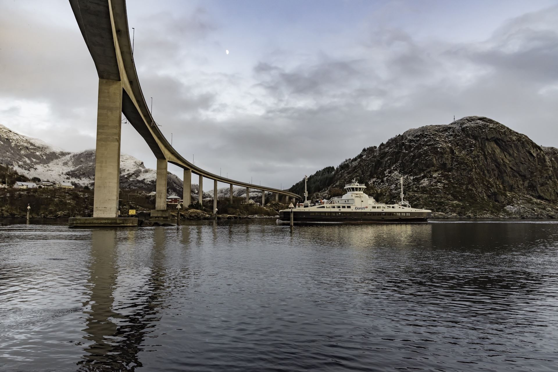 Curving concrete bridge crosses a calm fjord as a white ferry sits near rocky mountains under a cloudy sky..