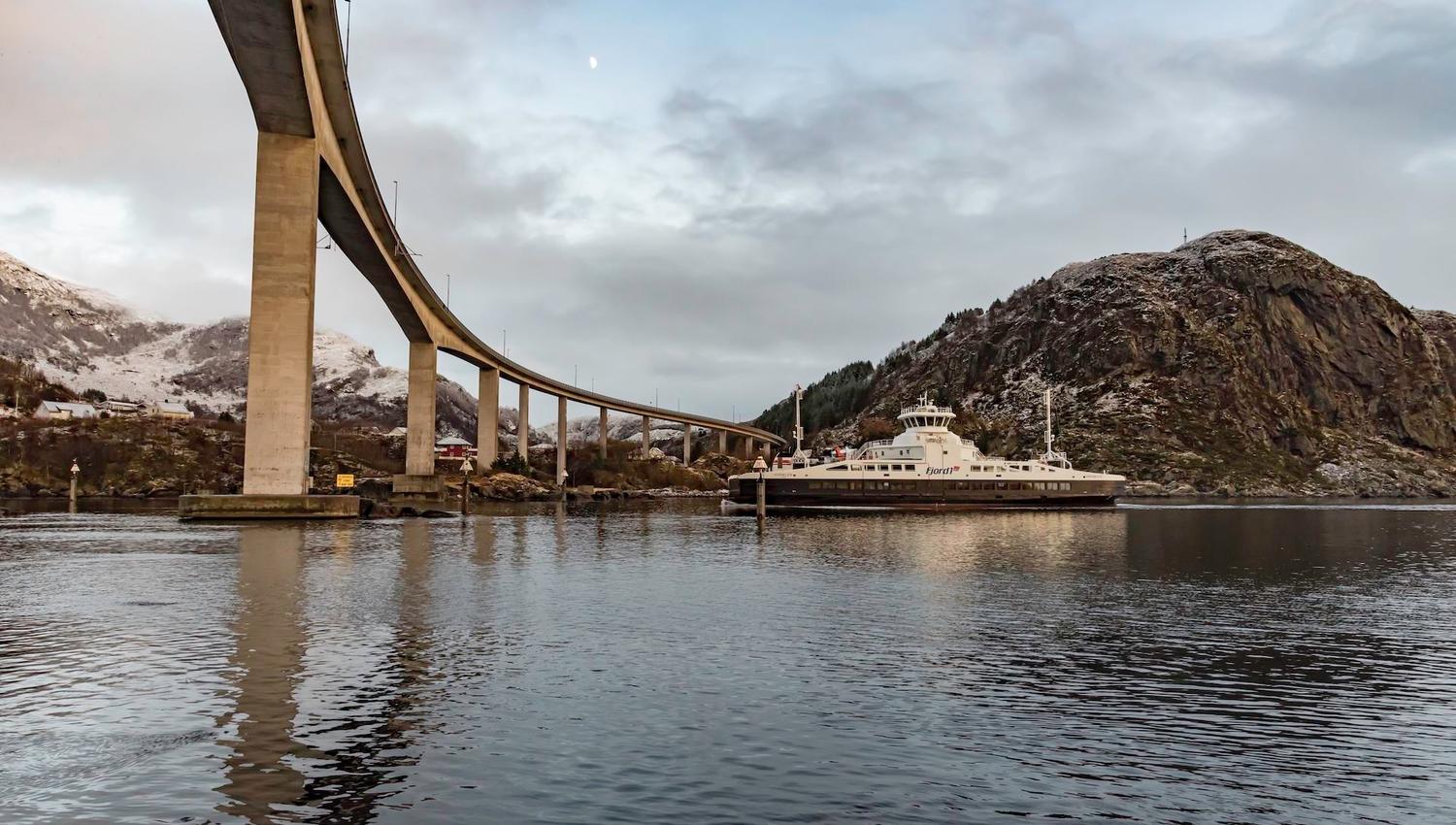 Curving concrete bridge crosses a calm fjord as a white ferry sits near rocky mountains under a cloudy sky..