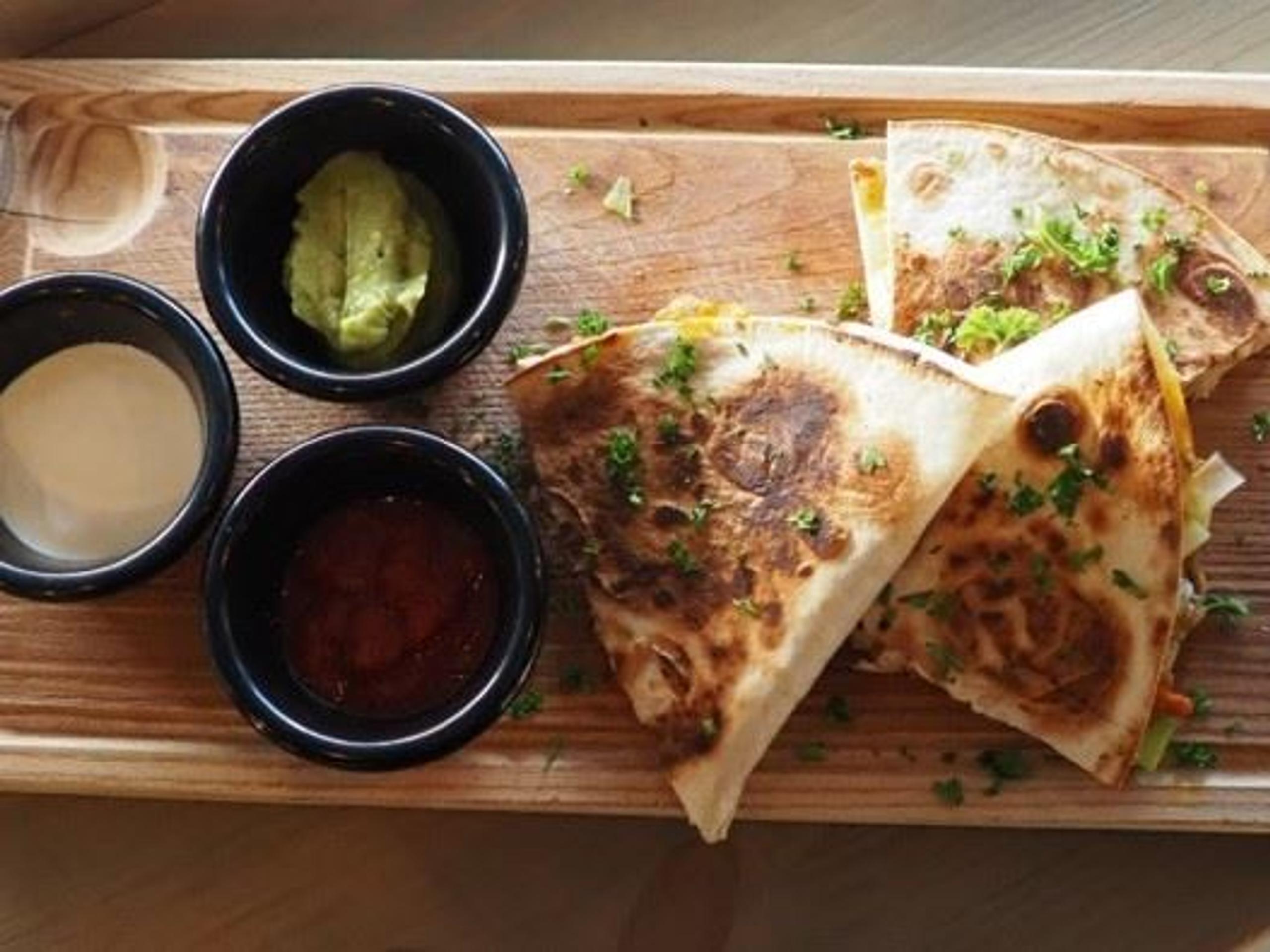Three toasted quesadilla wedges on a wooden board with small bowls of salsa, guacamole, and cream sauce.