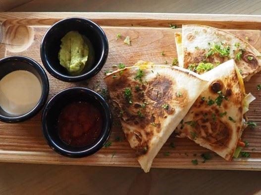 Three toasted quesadilla wedges on a wooden board with small bowls of salsa, guacamole, and cream sauce.