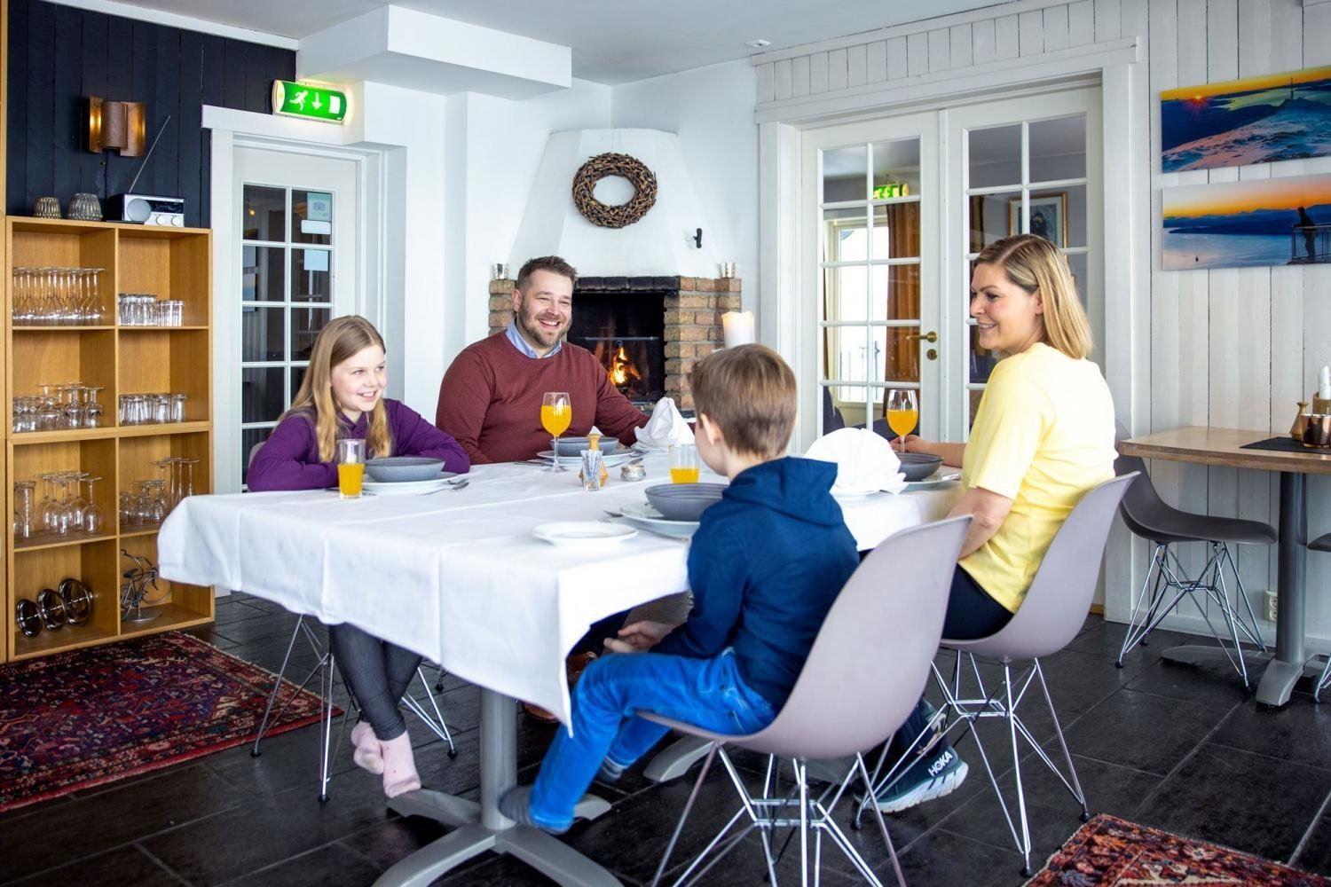 A family of four sits at a dining table with drinks and place settings in a bright room with a lit fireplace.