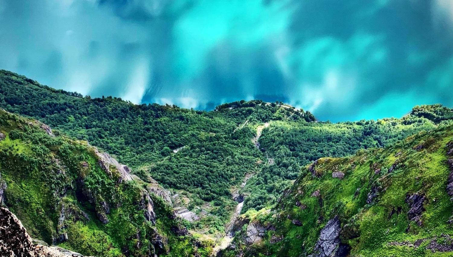 Steep green mountain slopes and rocky ravine descend toward a turquoise lake partly covered by low clouds..