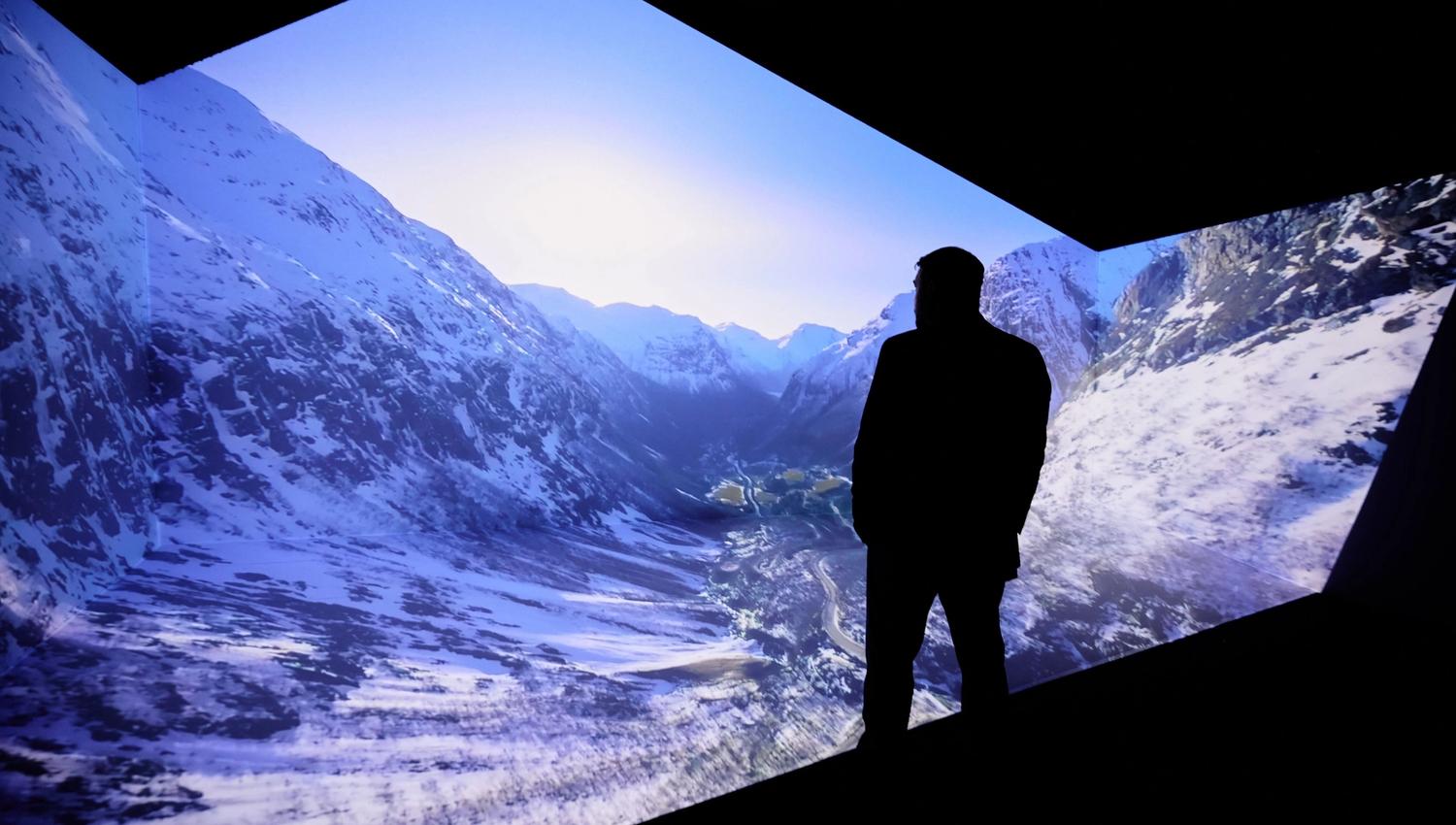 Ein person i silhuett står framfor ein stor projeksjon av snødekte fjell og dal under klar blå himmel..