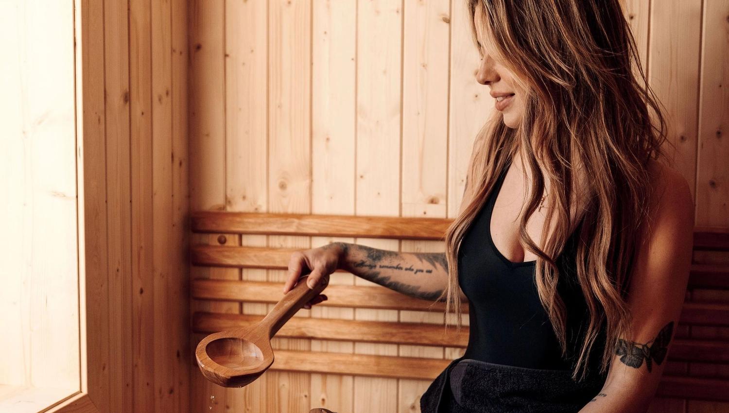 Woman sitting on a wooden sauna bench holding a ladle over a water bucket, with warm light and wood walls at the Seahouse at Hornelen Panorama in Bremanger..