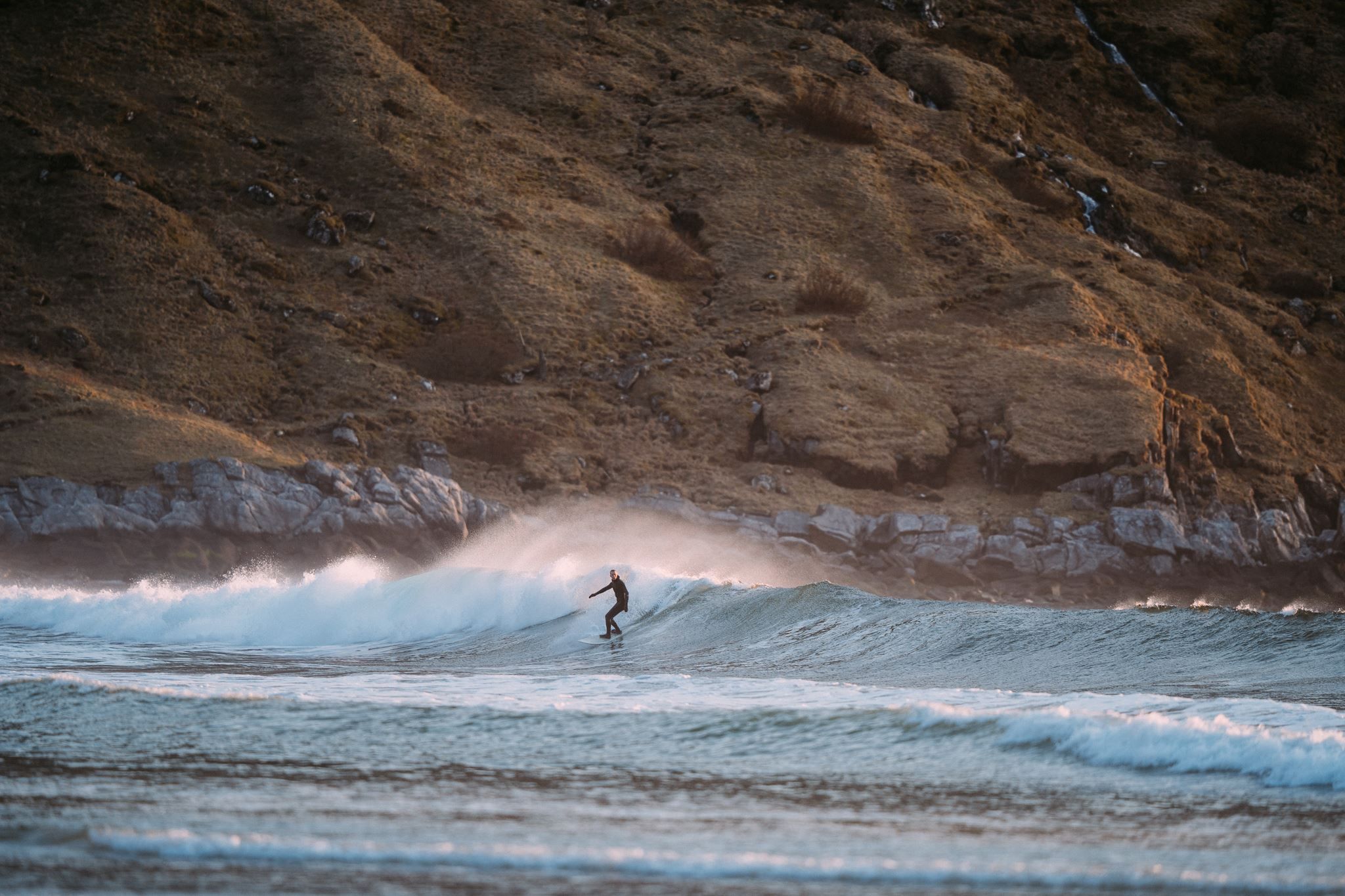 A surfer in a wetsuit rides a small breaking wave near a rocky shoreline with grassy hills in the background.