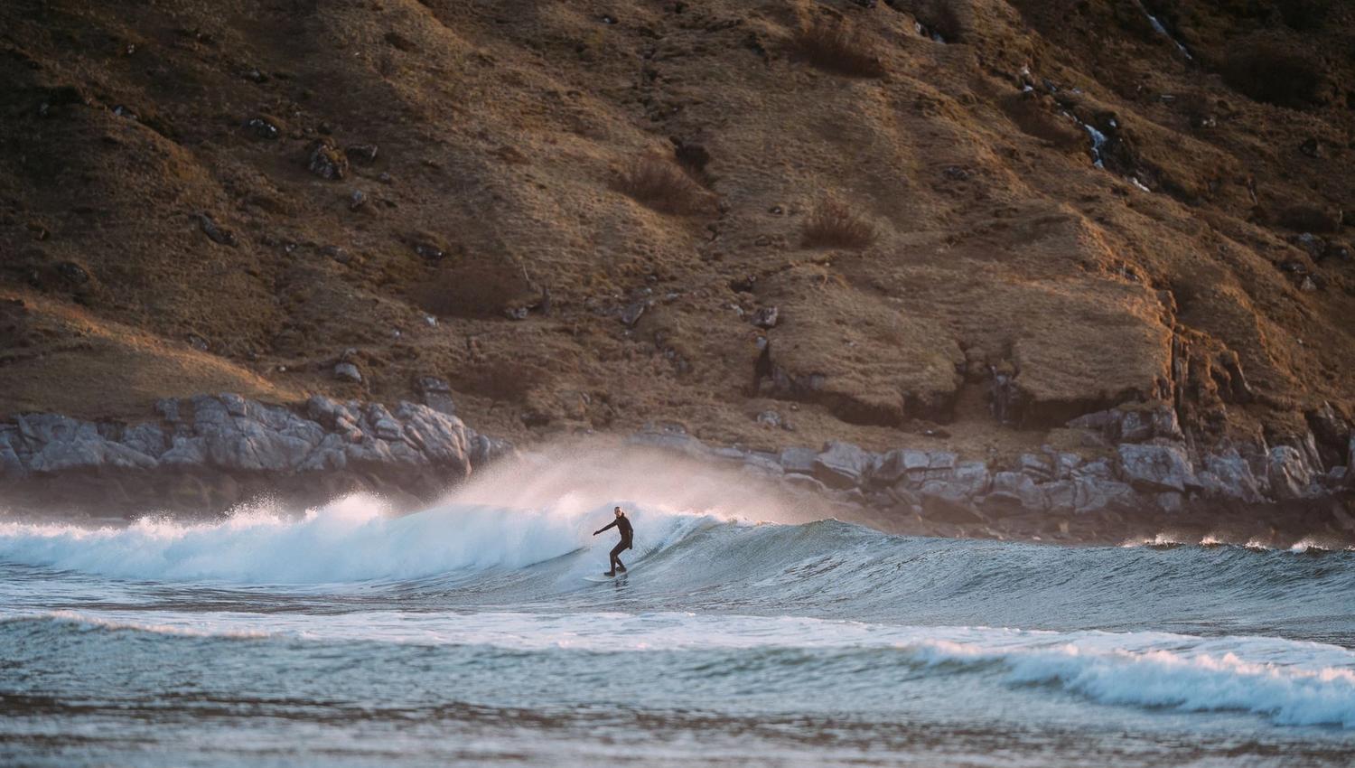 A surfer in a wetsuit rides a small breaking wave near a rocky shoreline with grassy hills in the background.