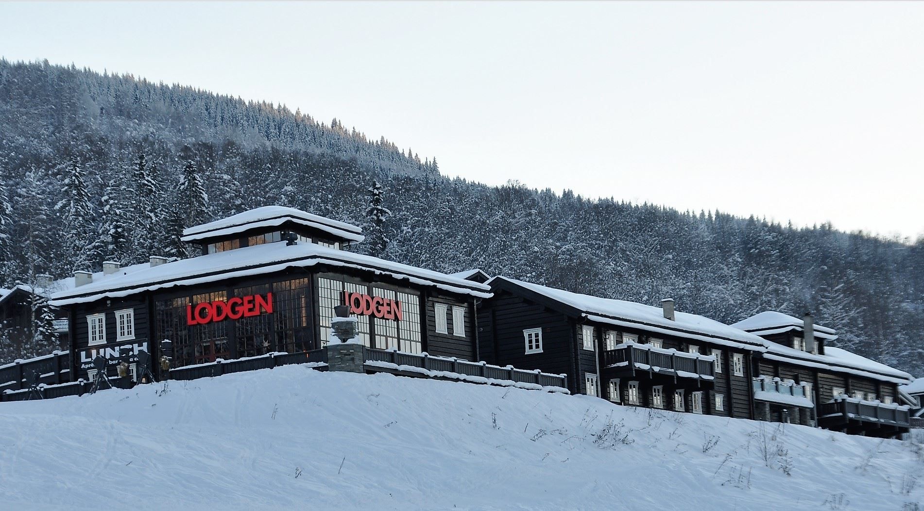 Snødekt, mørk trebygning med røde bokstaver på fasaden, foran en skogkledd fjellside i vinterlys..
