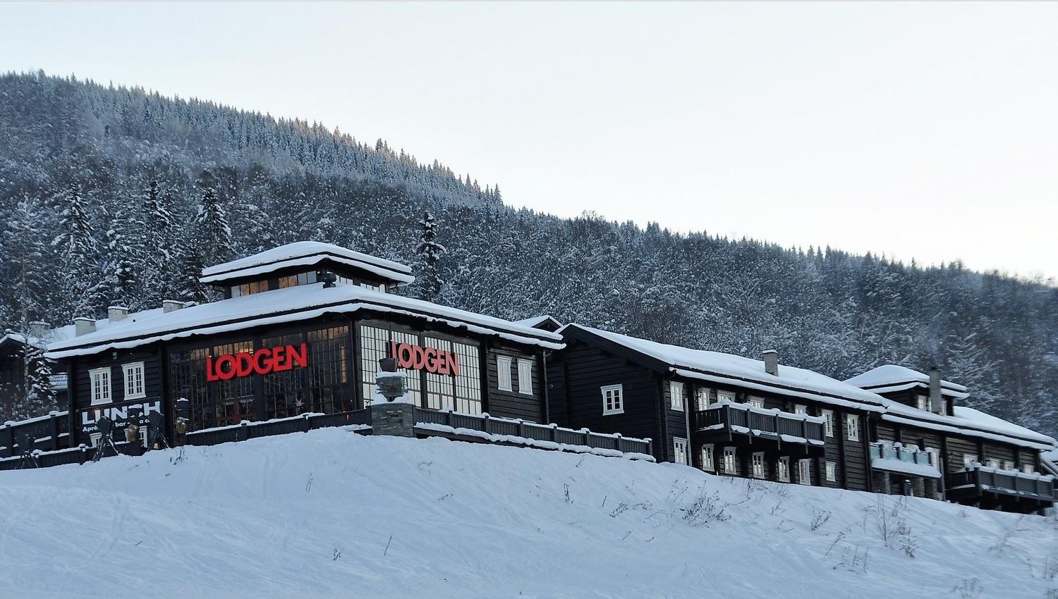 Snødekt, mørk trebygning med røde bokstaver på fasaden, foran en skogkledd fjellside i vinterlys..