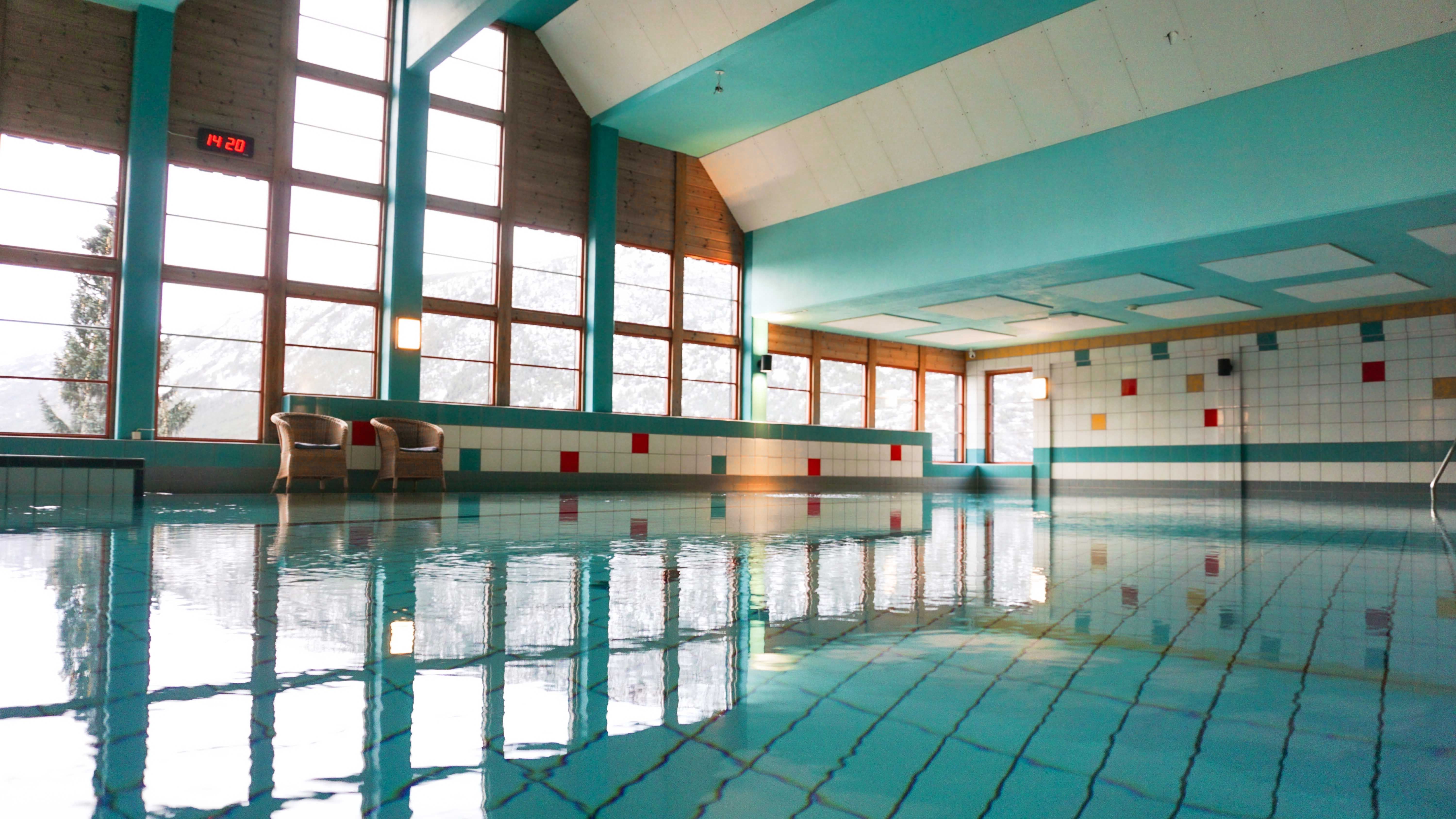 Quiet indoor swimming pool with turquoise walls, tiled deck, tall windows, and reflections on calm water..