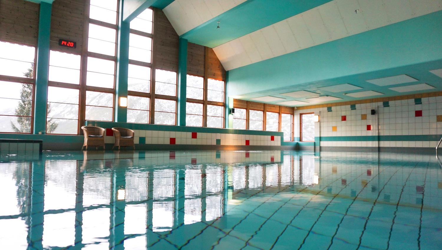 Quiet indoor swimming pool with turquoise walls, tiled deck, tall windows, and reflections on calm water..