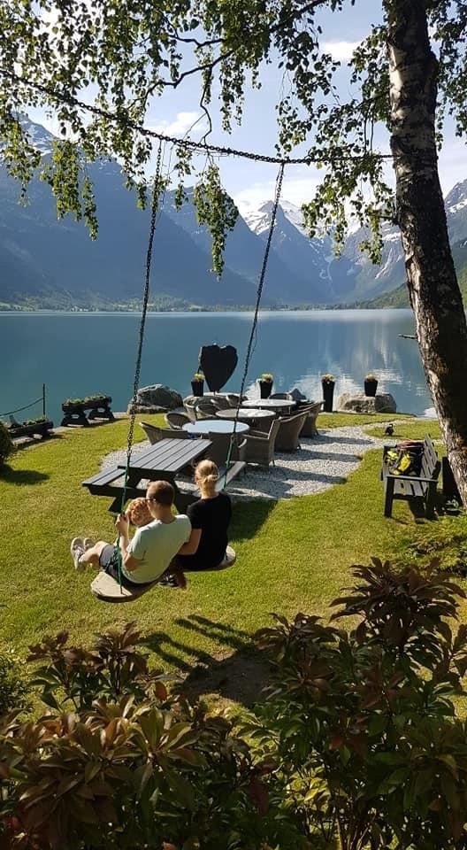 Two people sit on a swing in a grassy yard beside outdoor chairs and a calm lake with mountains in the distance.