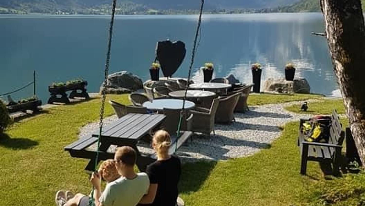 Two people sit on a swing in a grassy yard beside outdoor chairs and a calm lake with mountains in the distance.