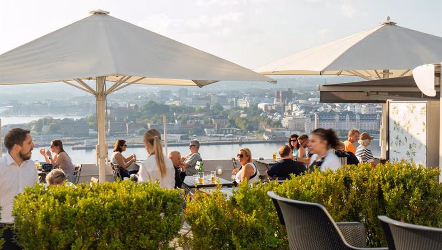 Guests enjoy food and drinks on a sunny rooftop terrace with panoramic views over Oslo and the fjord.