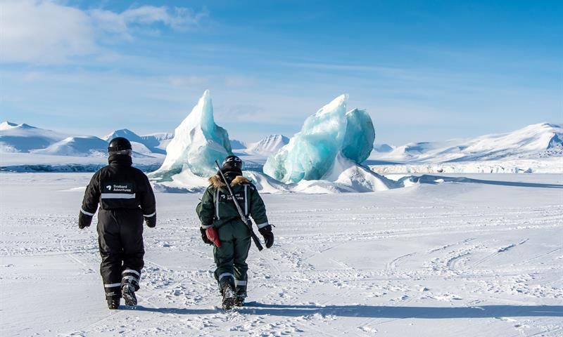 To personer går mot isfjell i snødekt landskap.