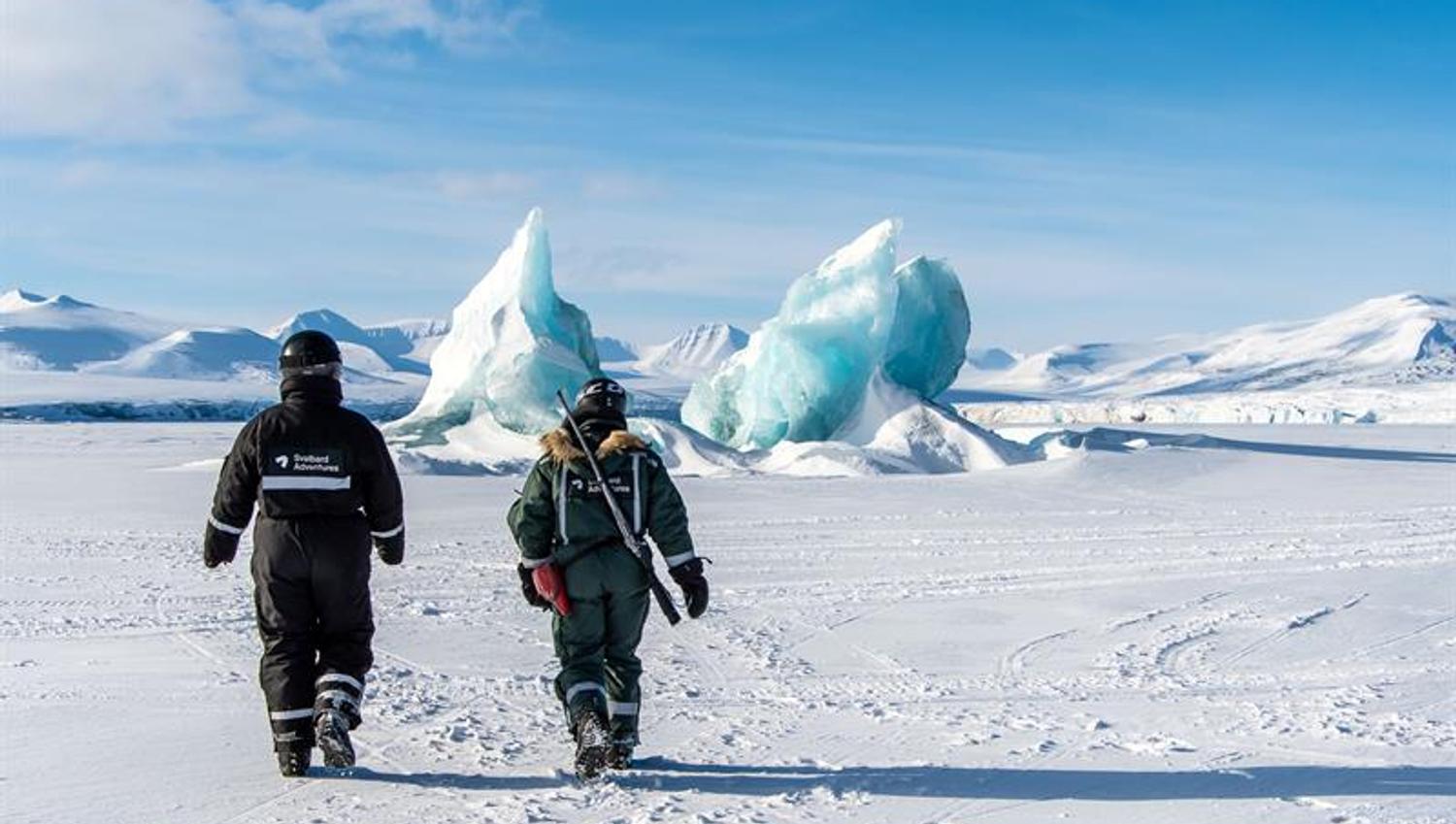 To personer går mot isfjell i snødekt landskap.