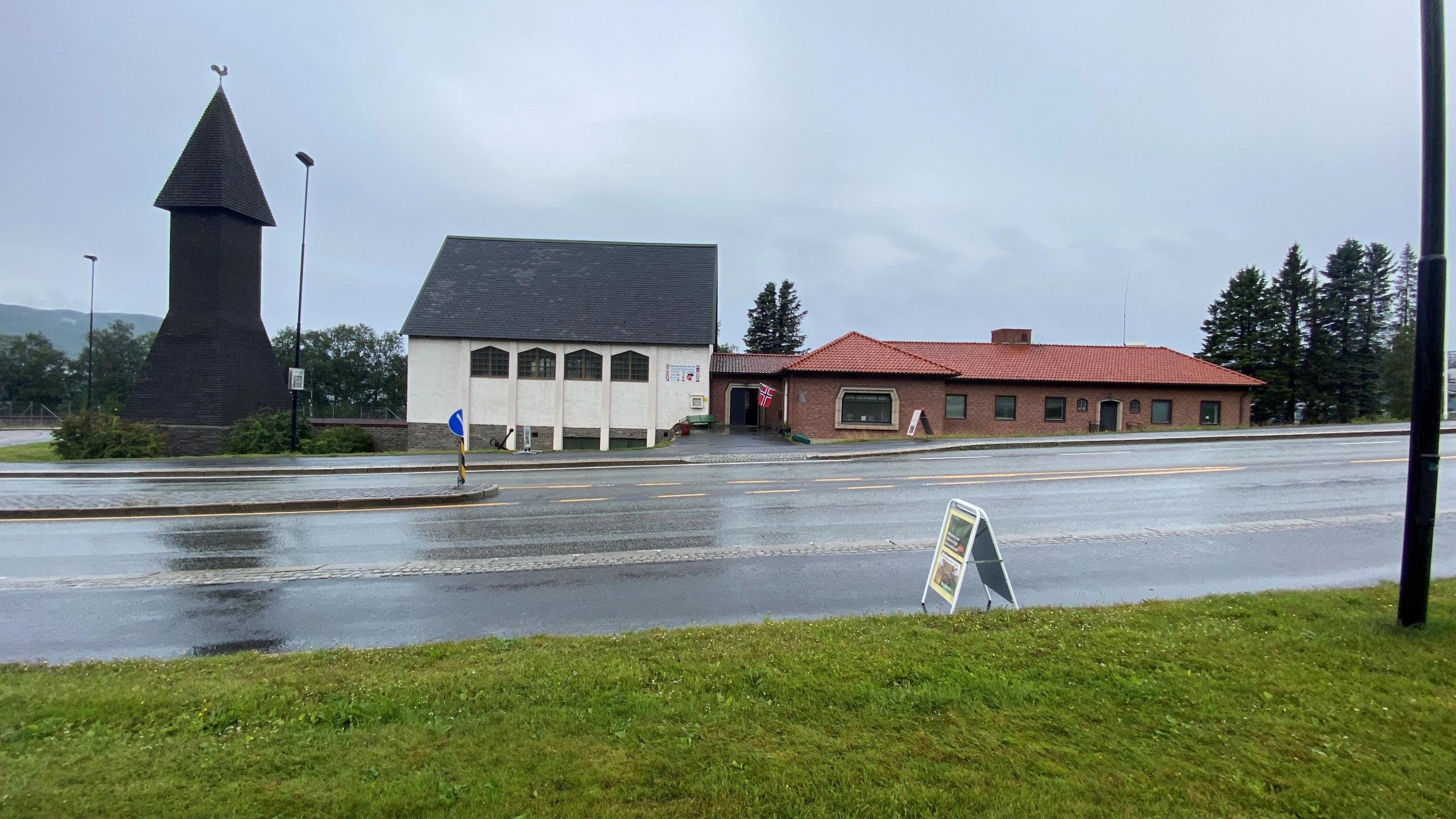 Wet roadway in front of a dark wooden tower and low buildings with a red roof, under an overcast sky.