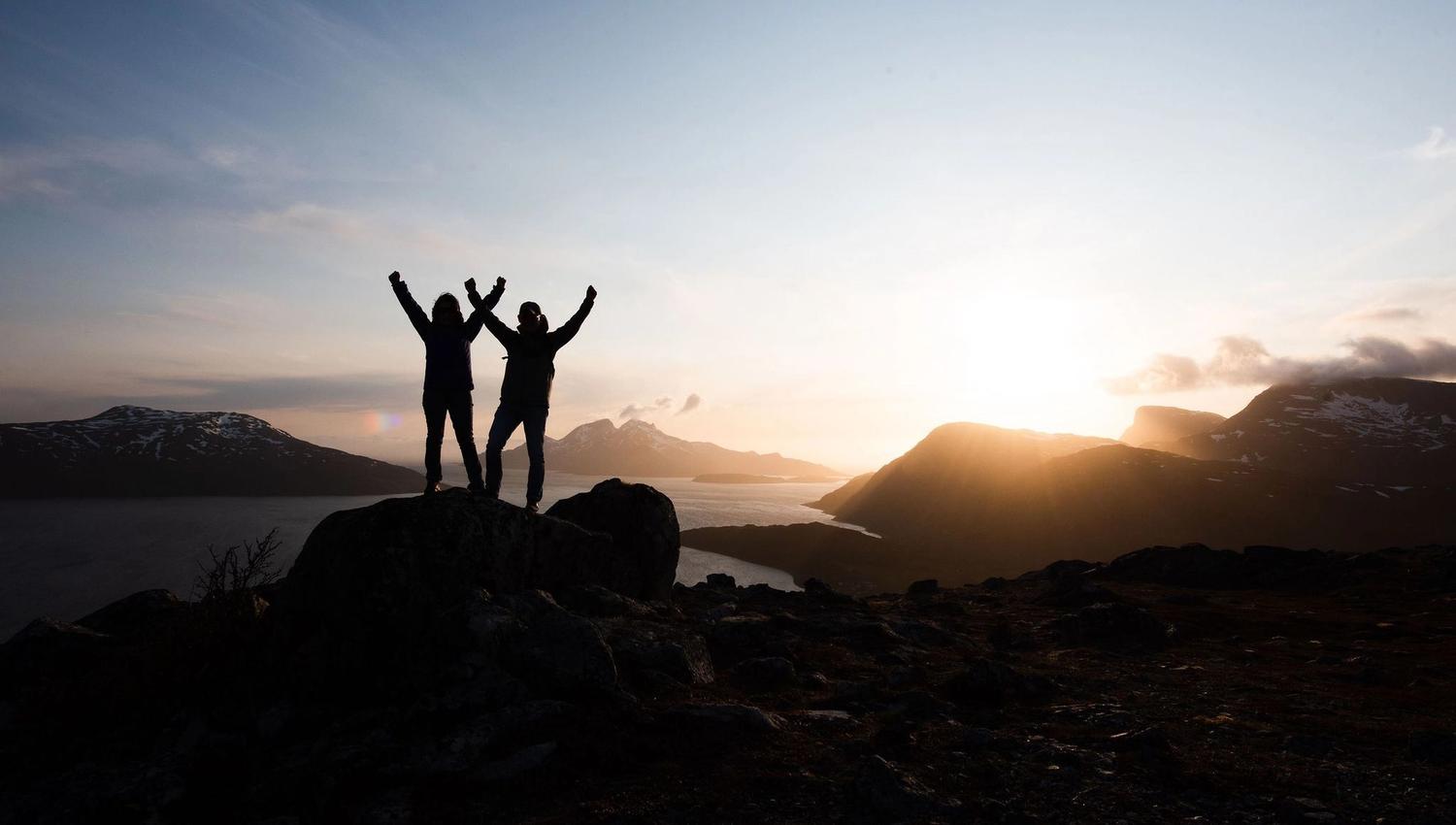 Two people raising their hands in the air in front of the midnight sun