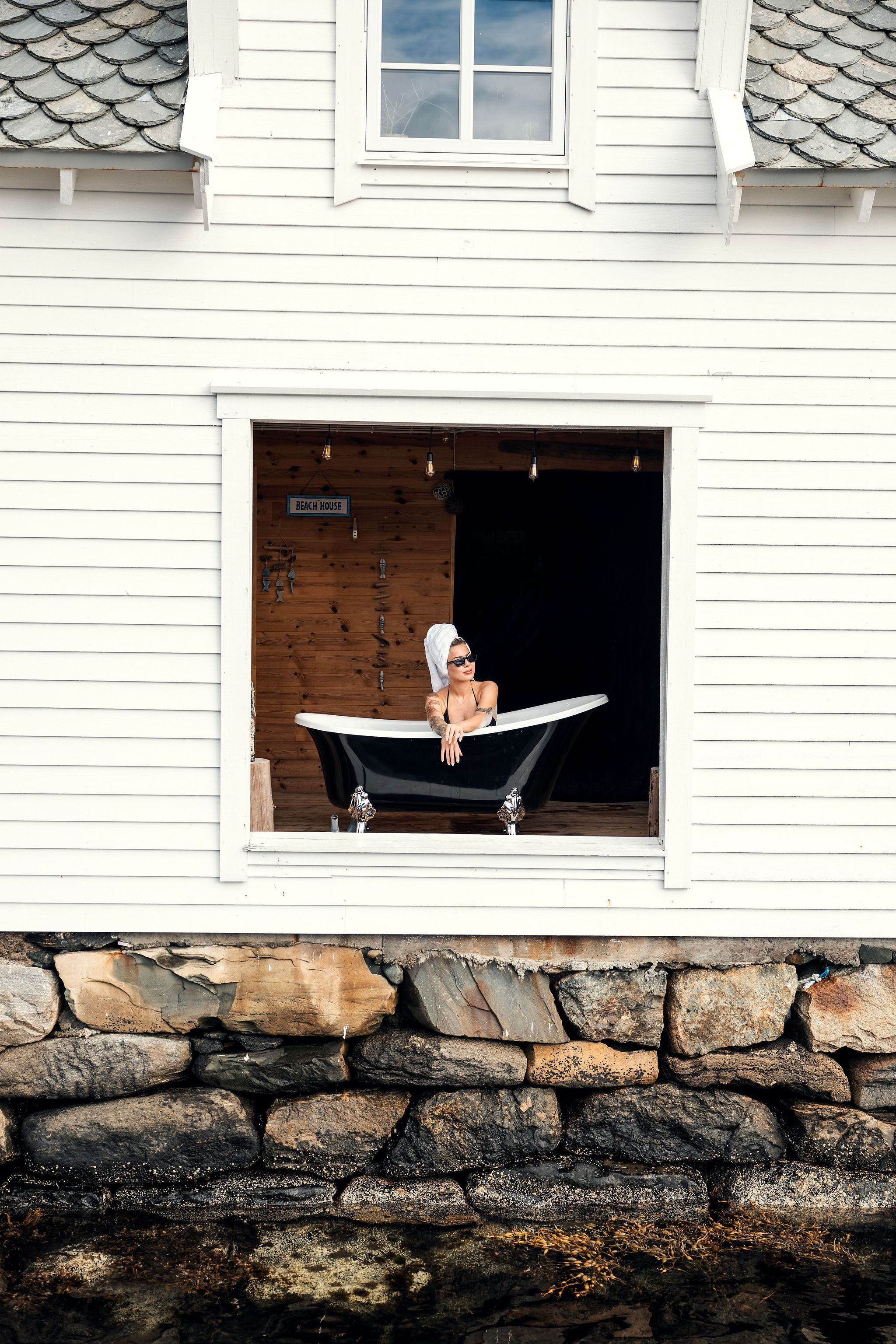 Person reclining in a black bathtub set inside a large window opening of a white beach house above a stone wall at the Seahouse at Hornelen Panorama in Bremanger..