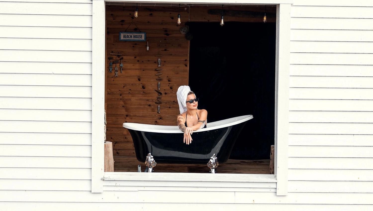 Person reclining in a black bathtub set inside a large window opening of a white beach house above a stone wall at the Seahouse at Hornelen Panorama in Bremanger..