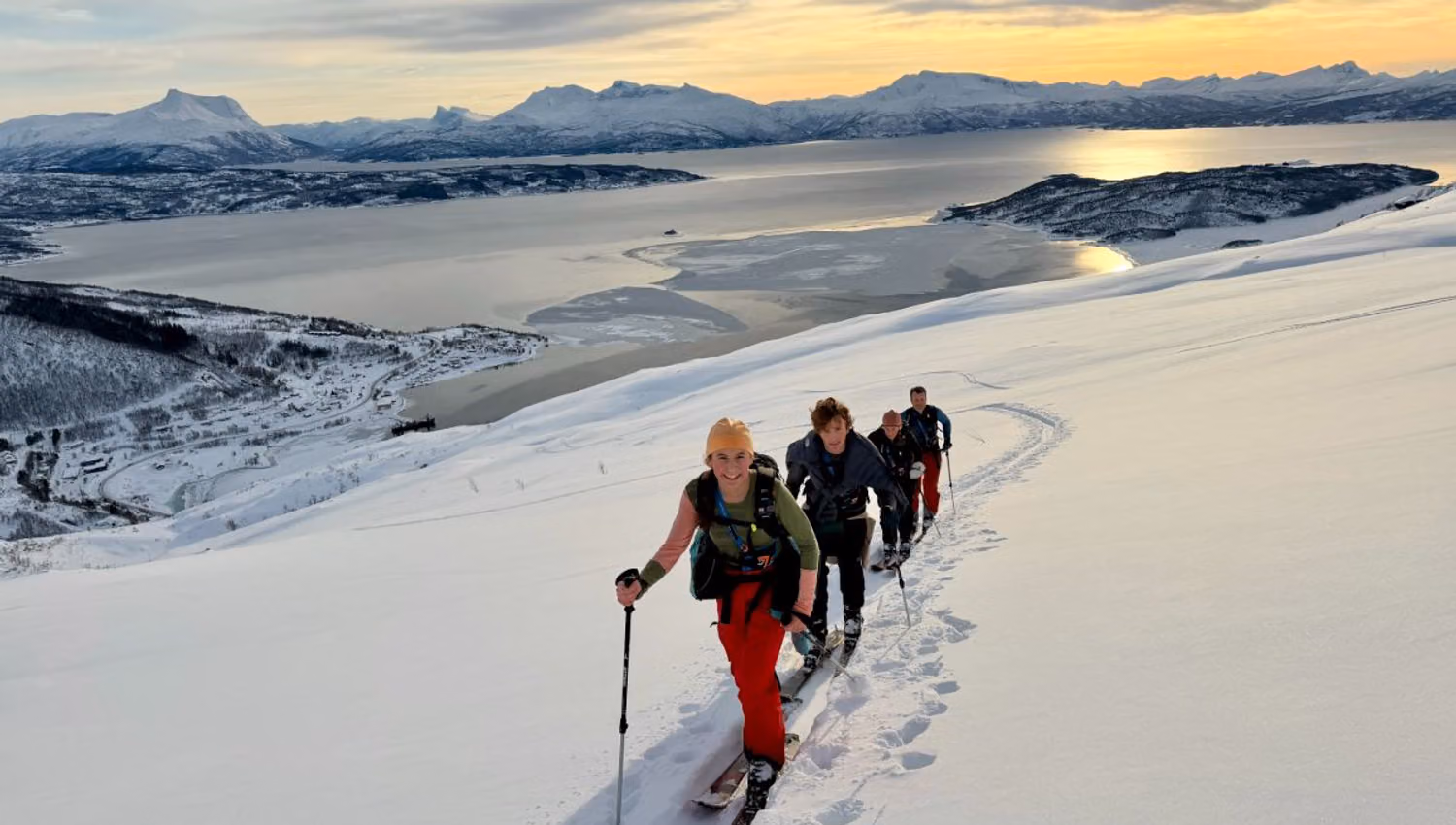 Four backcountry skiers climb a snowy ridge toward the camera, with a fjord and mountains under a low sun..